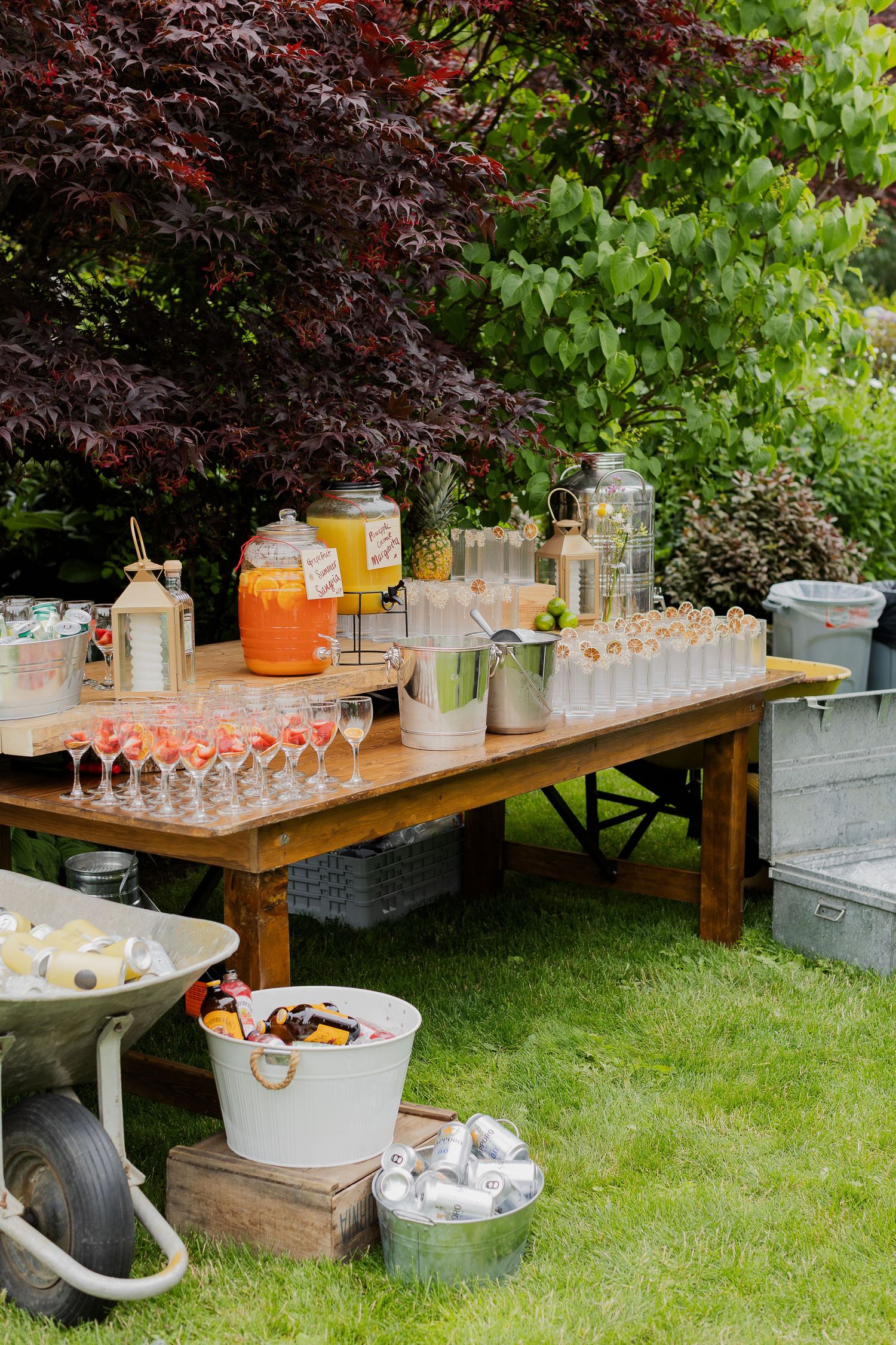 Outdoor wedding drink station with colorful beverages, champagne flutes, and ice buckets on a rustic wooden table.