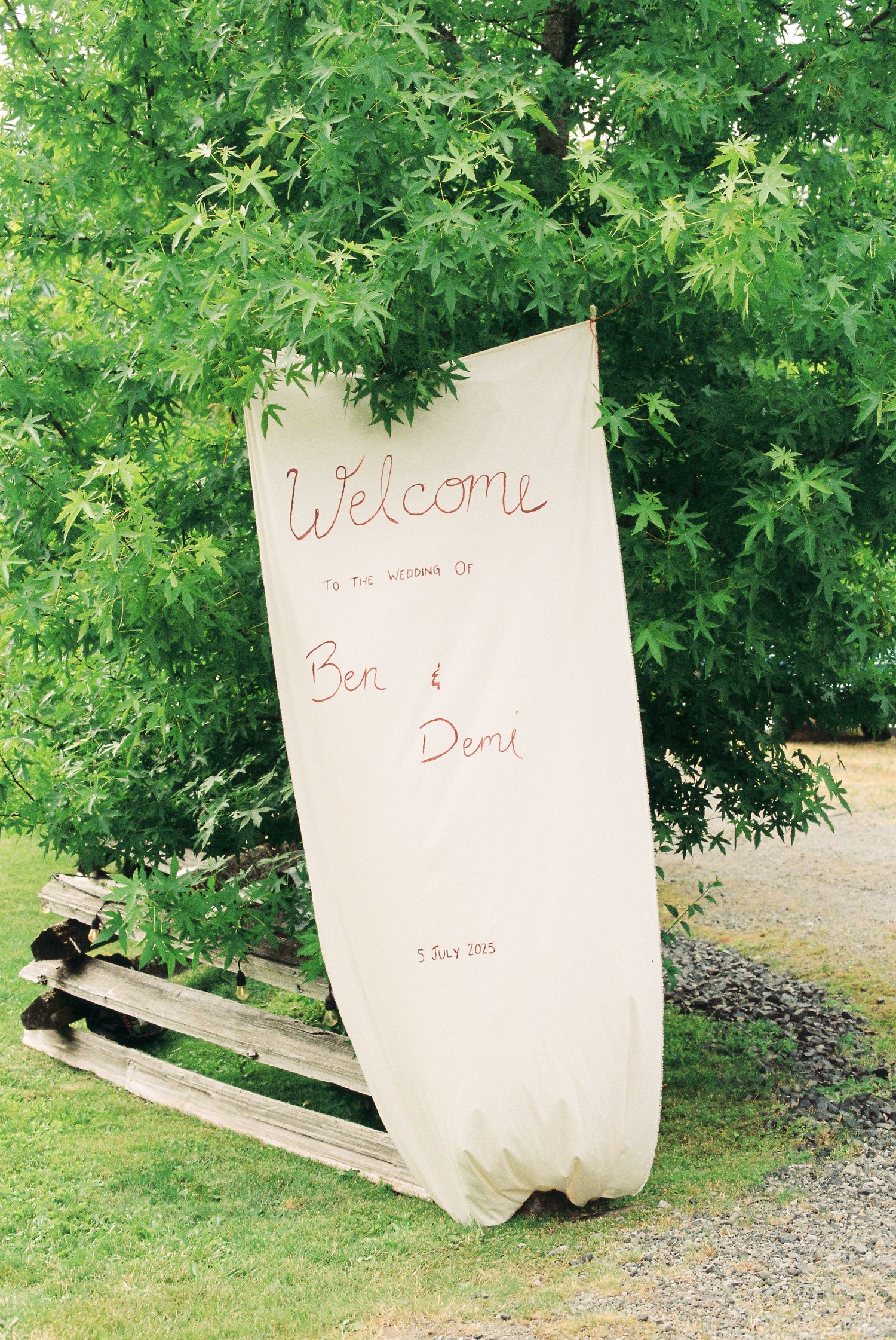 Handmade fabric welcome sign for Ben and Demi wedding hanging on wooden fence beneath maple tree.