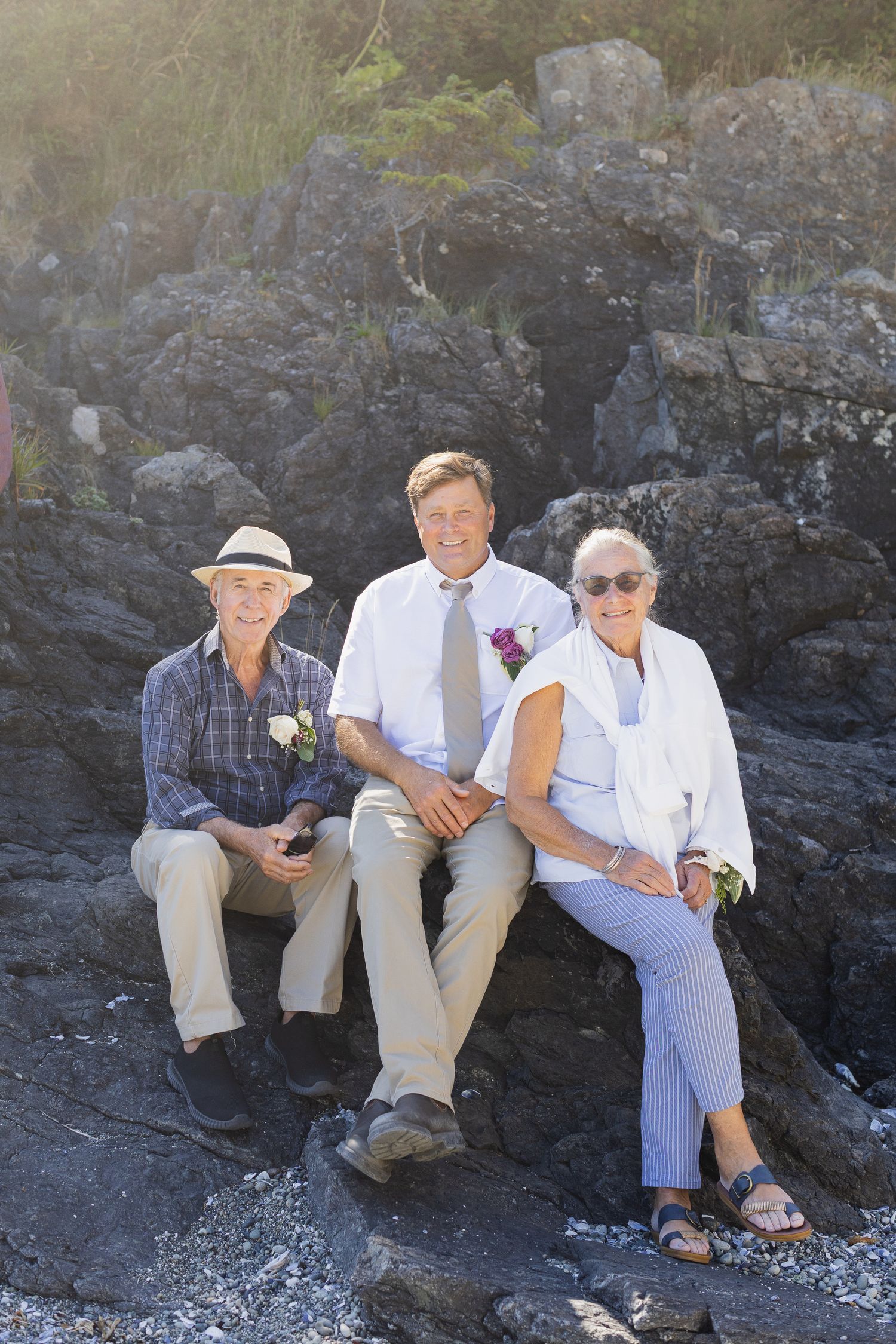 Groom sits between elderly parents on rocky beach during outdoor wedding celebration in sunlight.