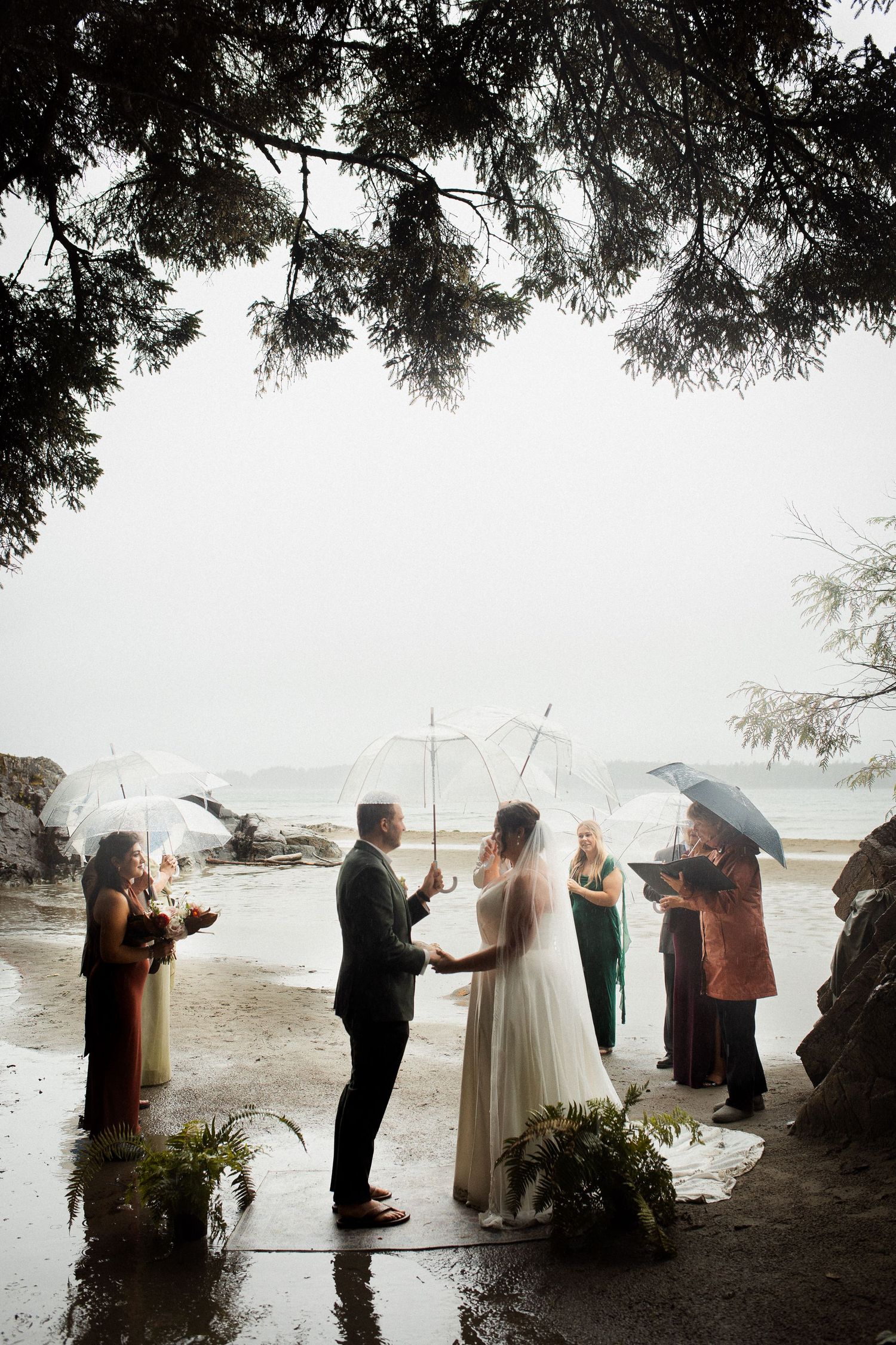 Bride and groom hold hands during intimate rainy beach wedding ceremony with clear umbrellas.