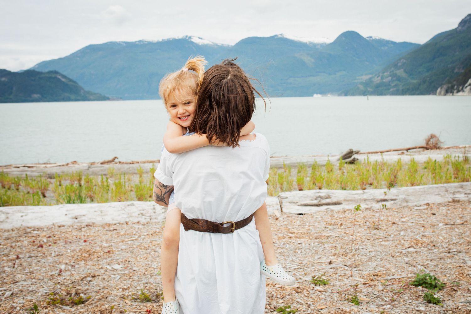 Mother carrying young daughter on her back near rocky shoreline with mountain lake views.