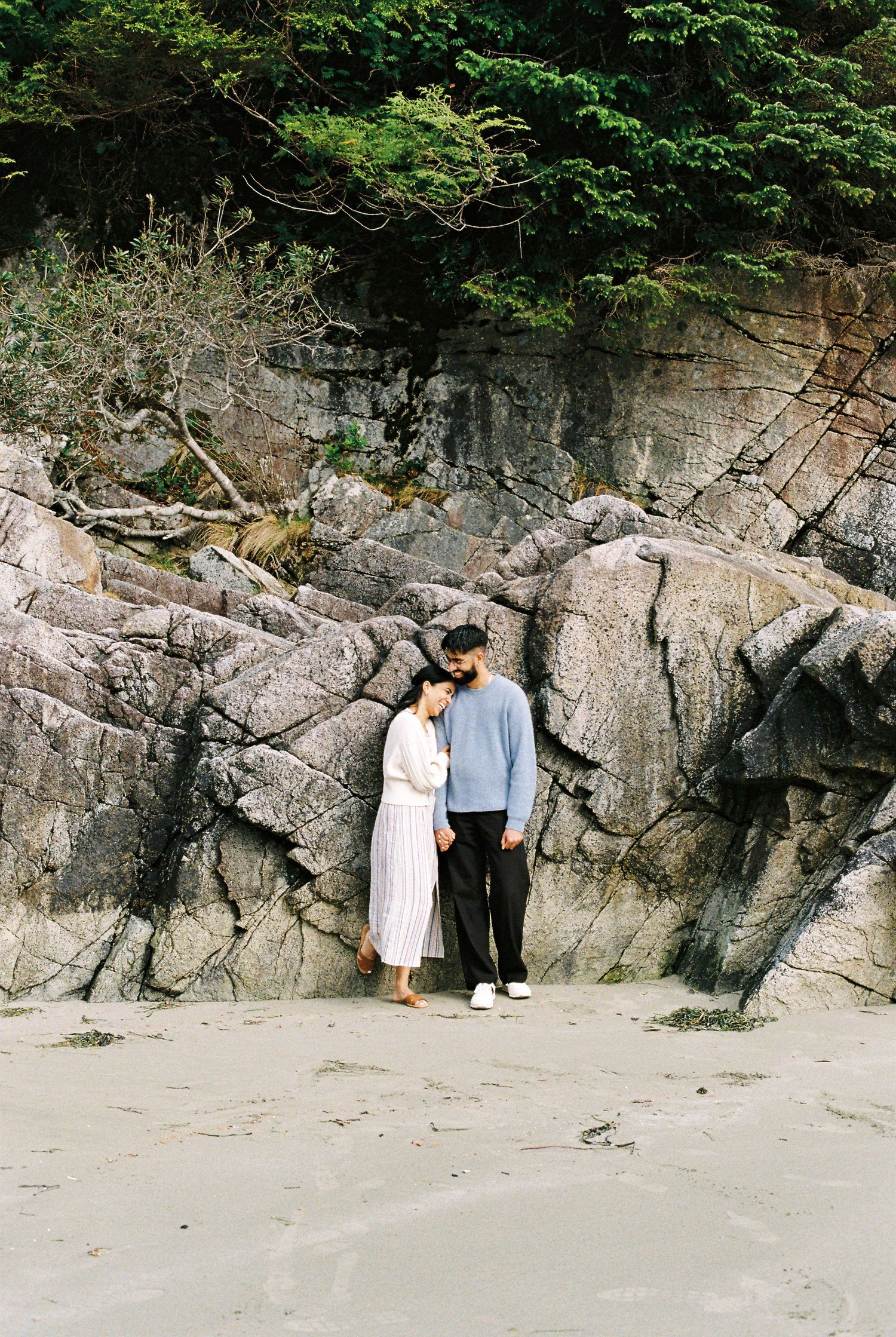 Couple standing close together against dramatic rock face on a sandy beach.