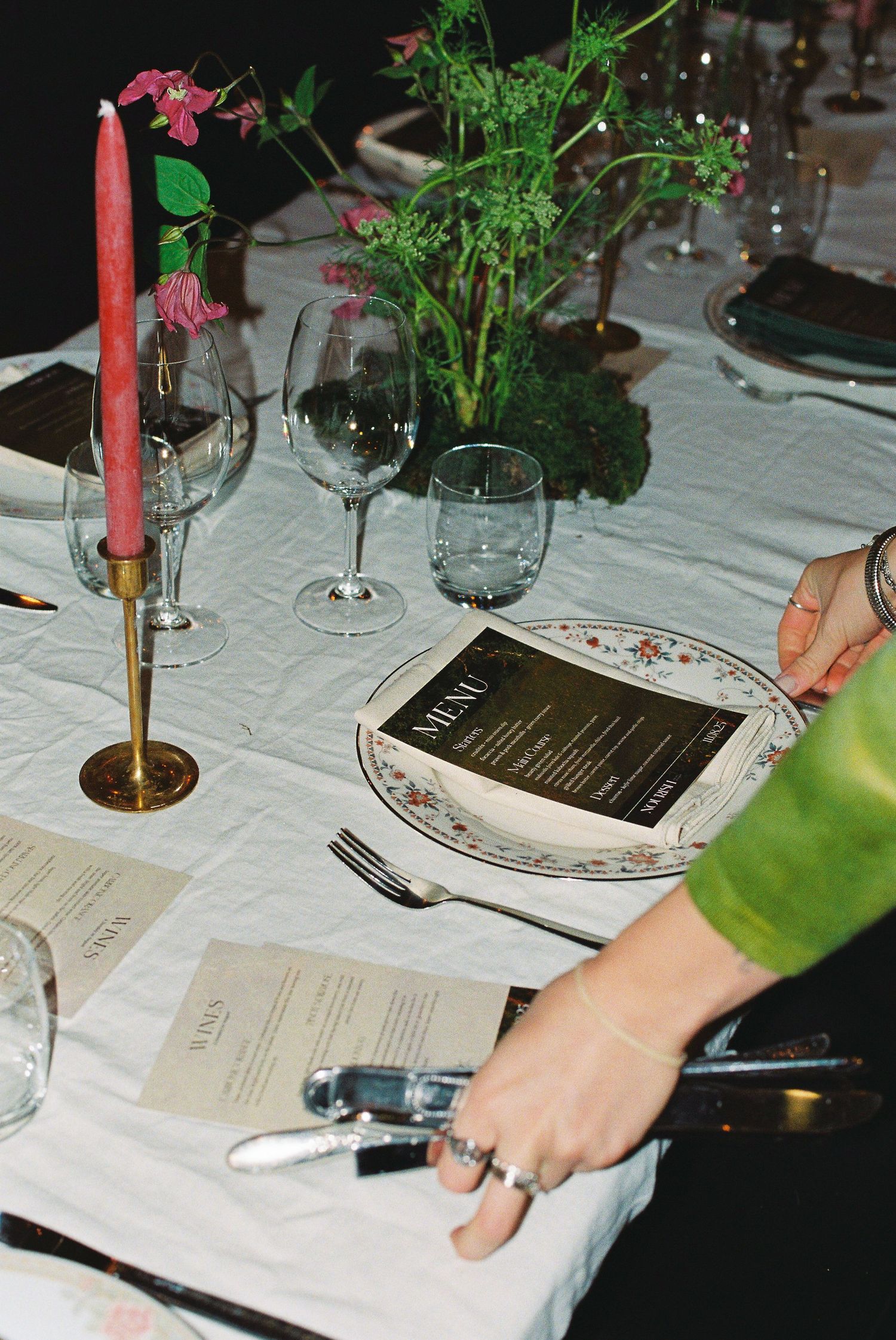 Elegant dinner table setting with pink taper candle, wine glasses, floral plates, and black menu card.