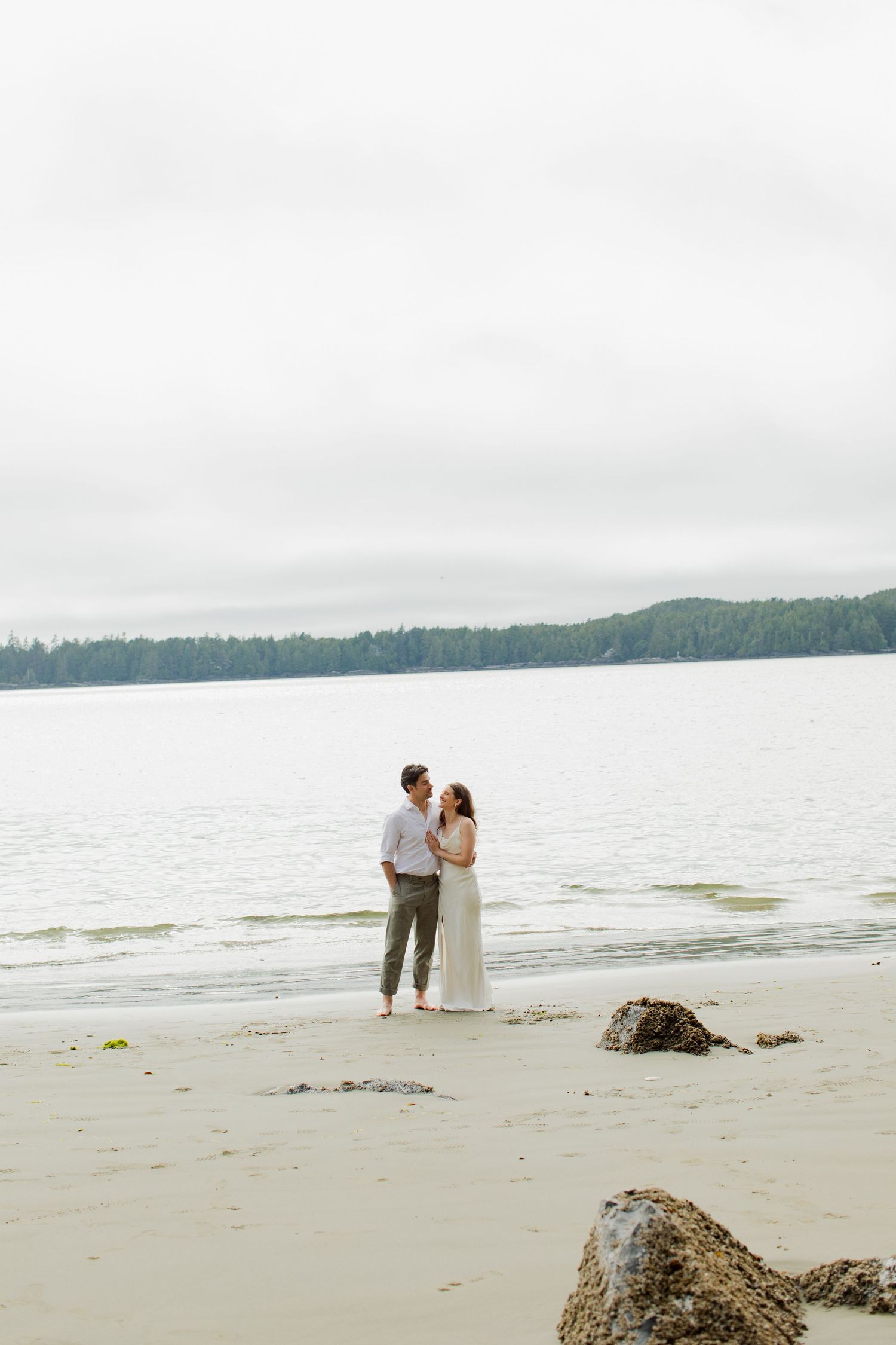 Couple embraces barefoot on a sandy beach shore, gazing at each other with calm ocean and trees behind.