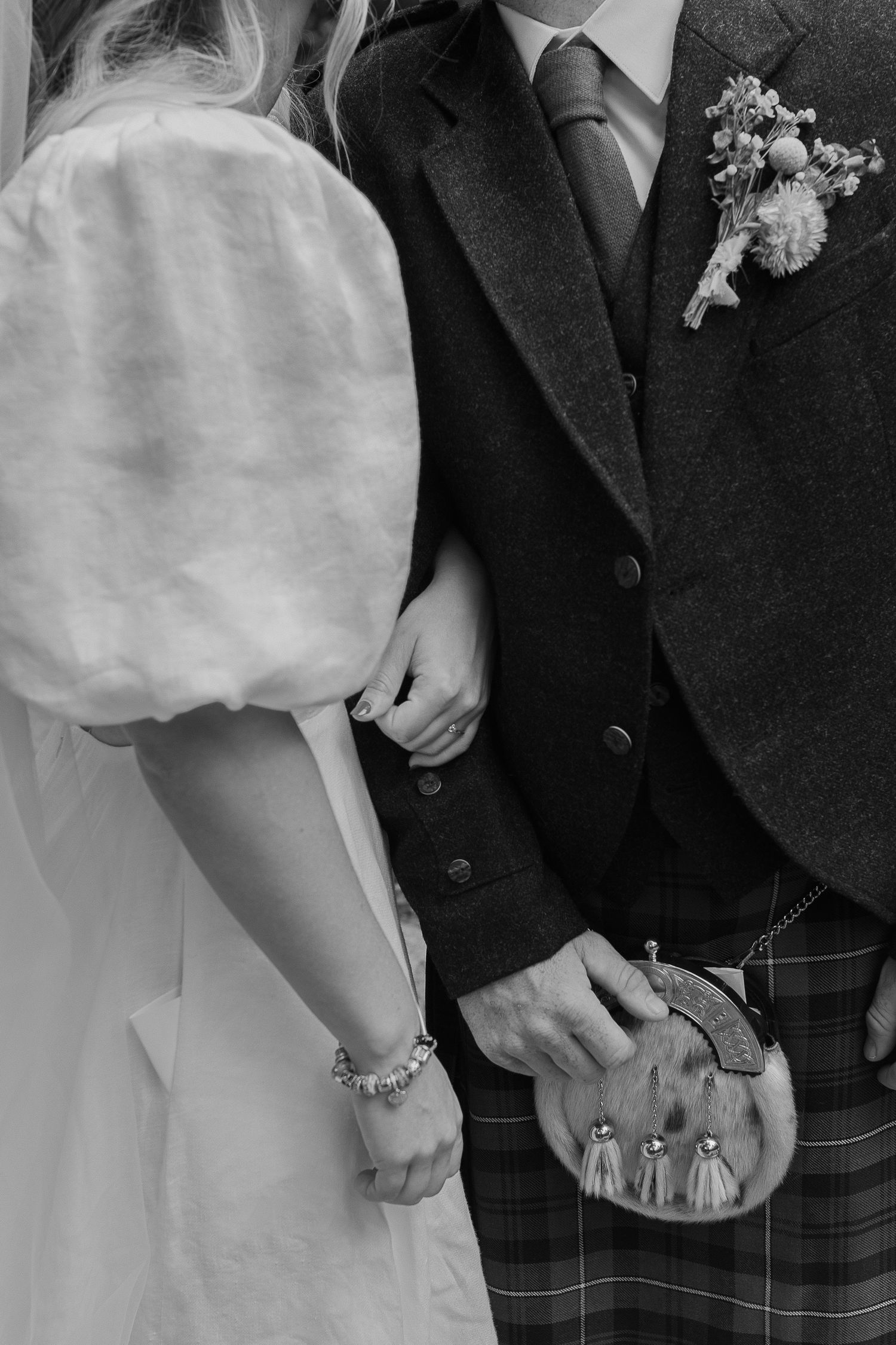 Black and white close-up of bride and groom in kilt holding hands at wedding.