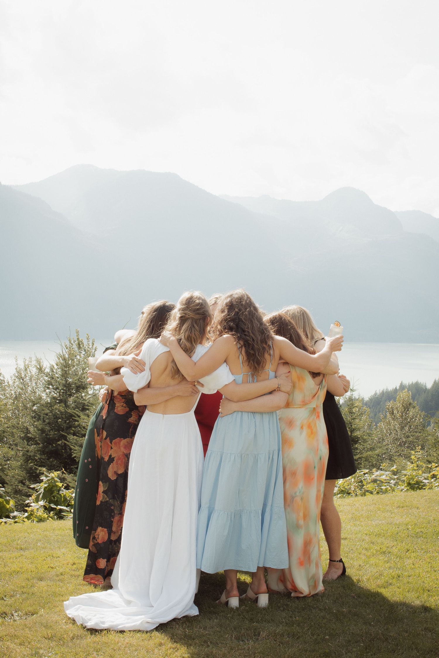 Bride and bridesmaids sharing group hug on lawn with stunning mountain lake view behind them.