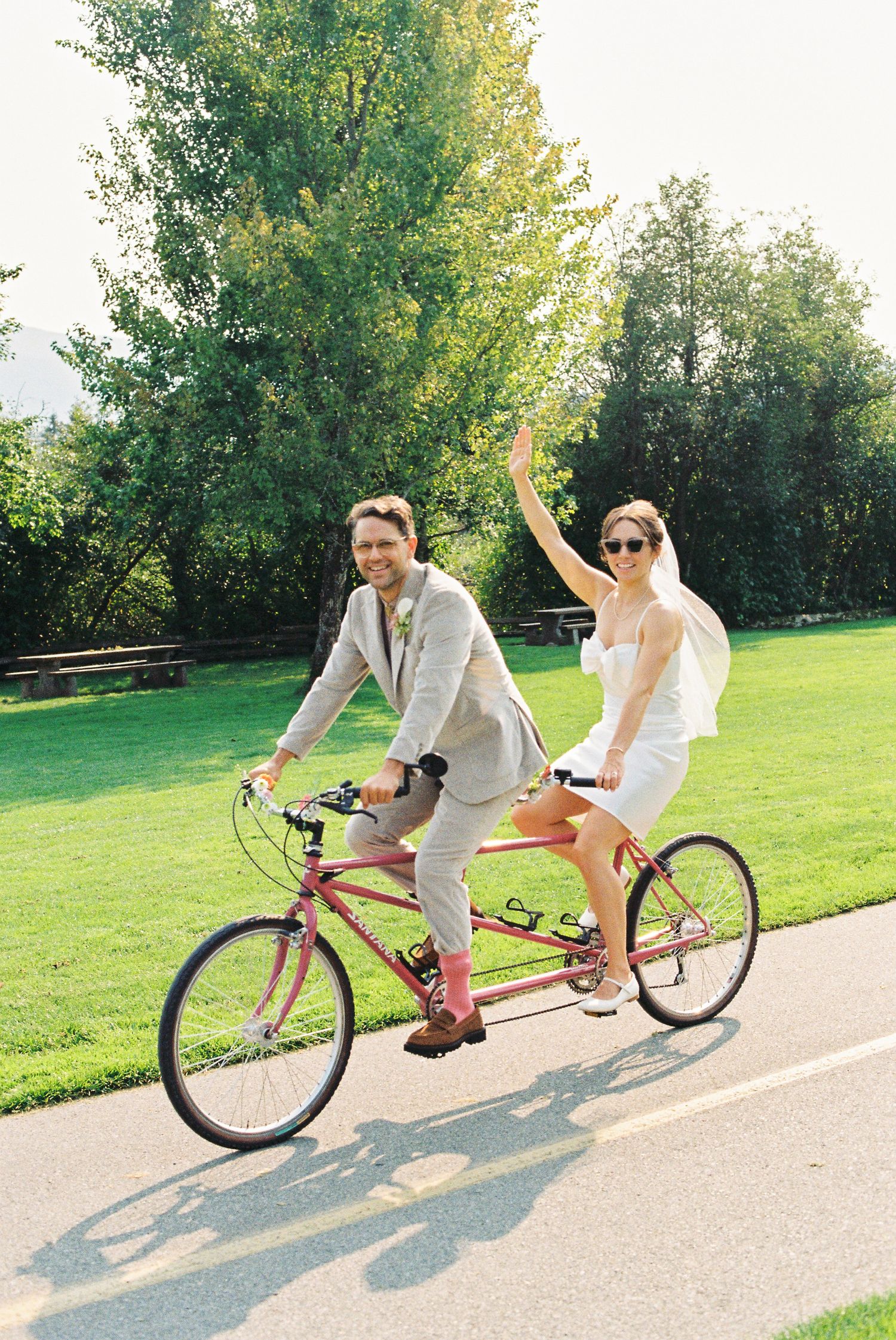 Bride and groom riding a red tandem bicycle through a sunny park, waving joyfully.