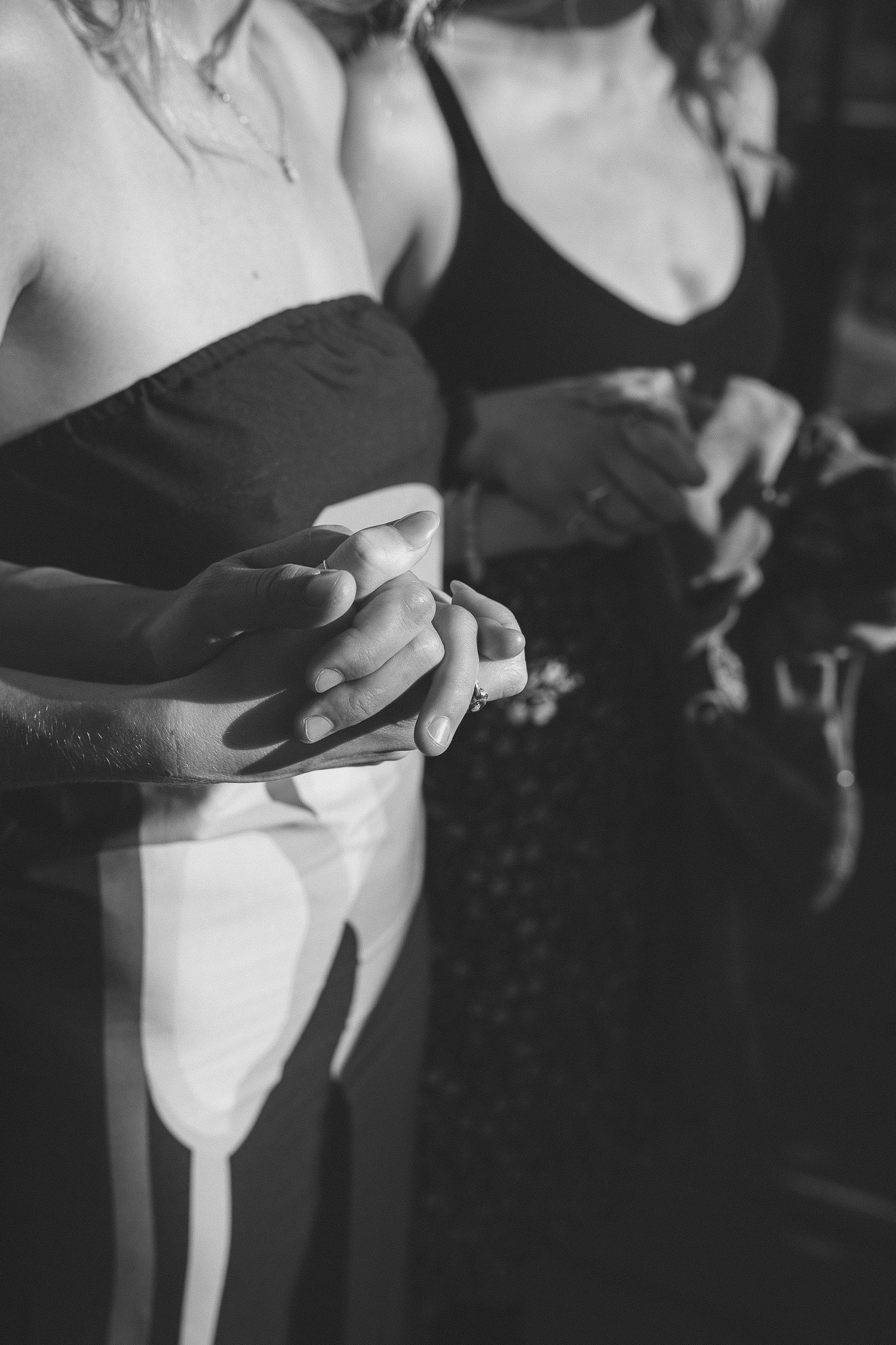 Black and white close-up of bridesmaids holding hands in black dresses during wedding ceremony.