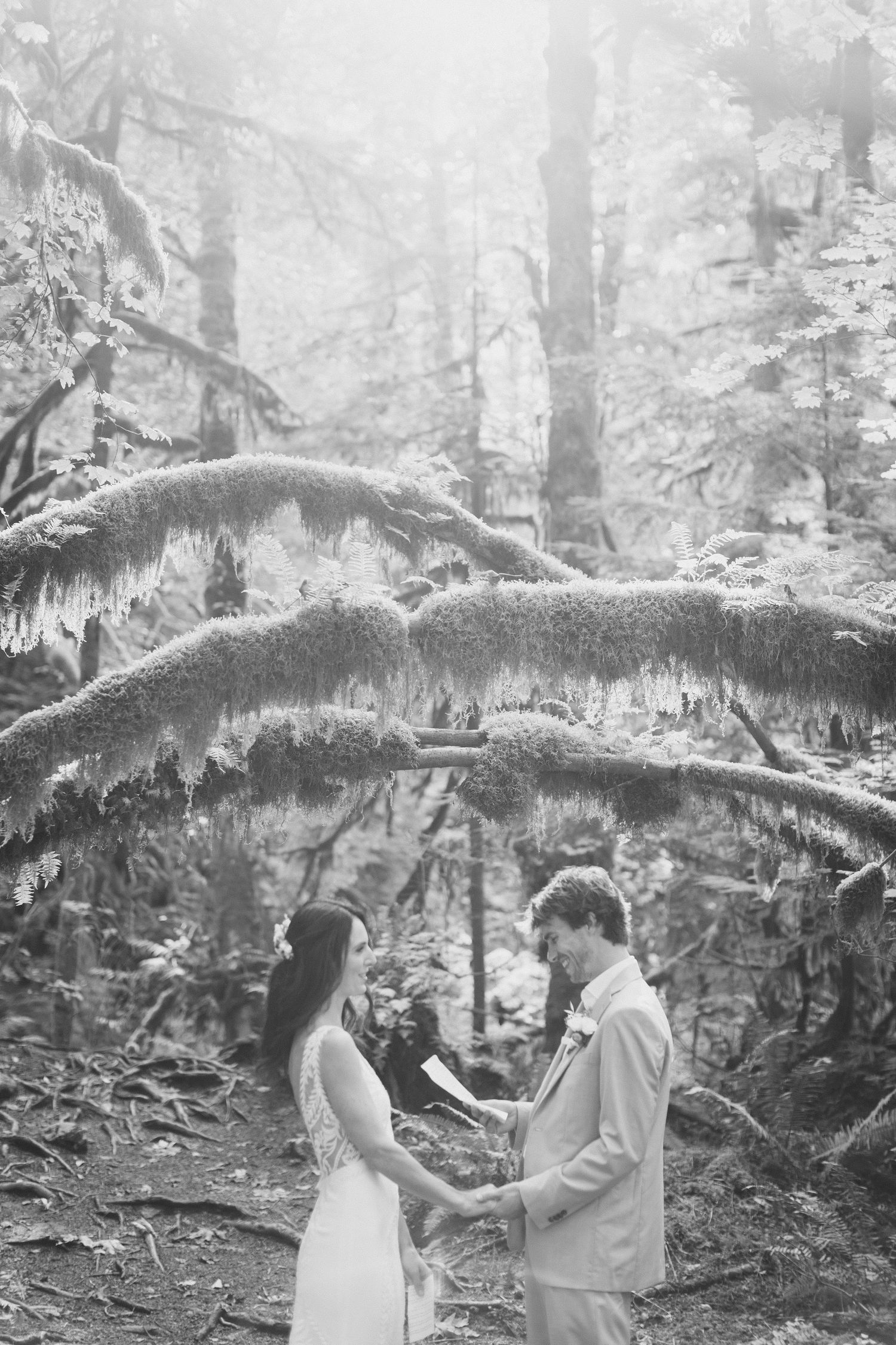 Black and white photo of couple exchanging vows in enchanted moss-covered forest setting.