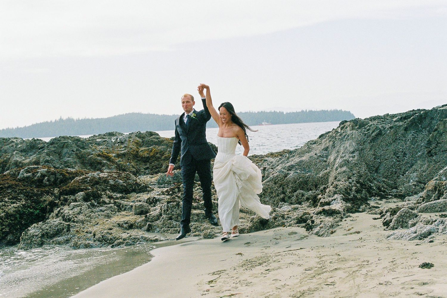 Joyful bride and groom celebrate on a rocky beach, raising hands in triumph after wedding.