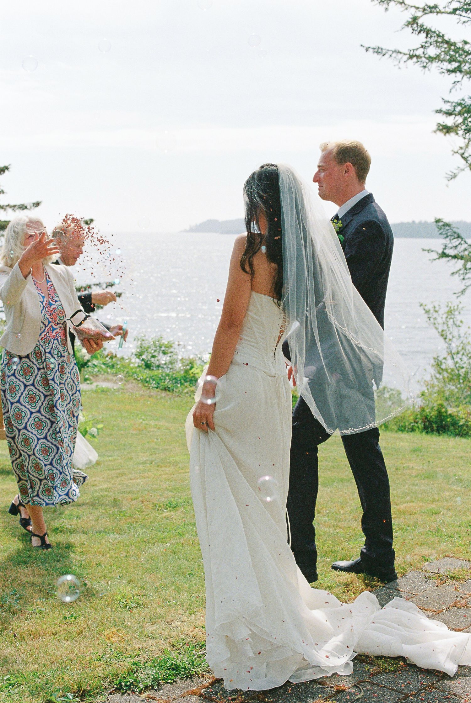 Guests toss confetti over bride in white veil and groom beside scenic ocean waterfront lawn.