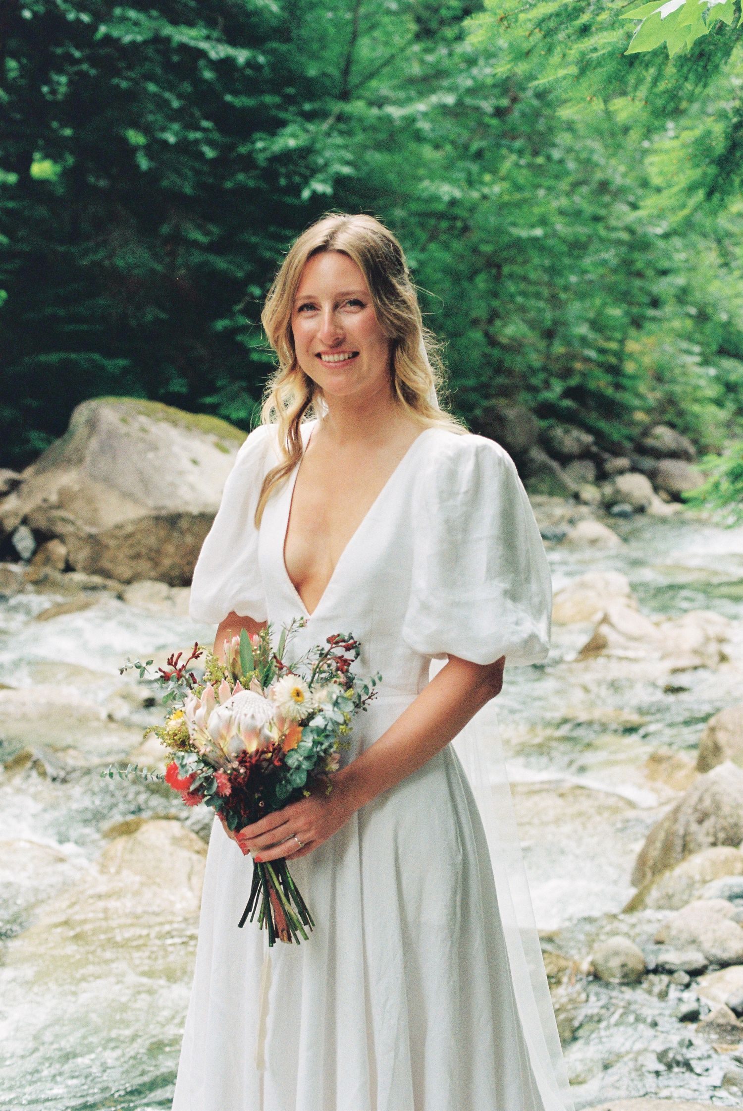 Smiling bride in white V-neck puff sleeve gown holds colorful wildflower bouquet by a creek.