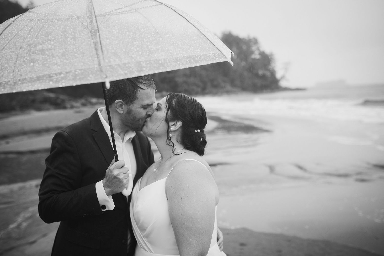 Black and white photo of a bride and groom kissing on a beach in the rain under a clear umbrella.