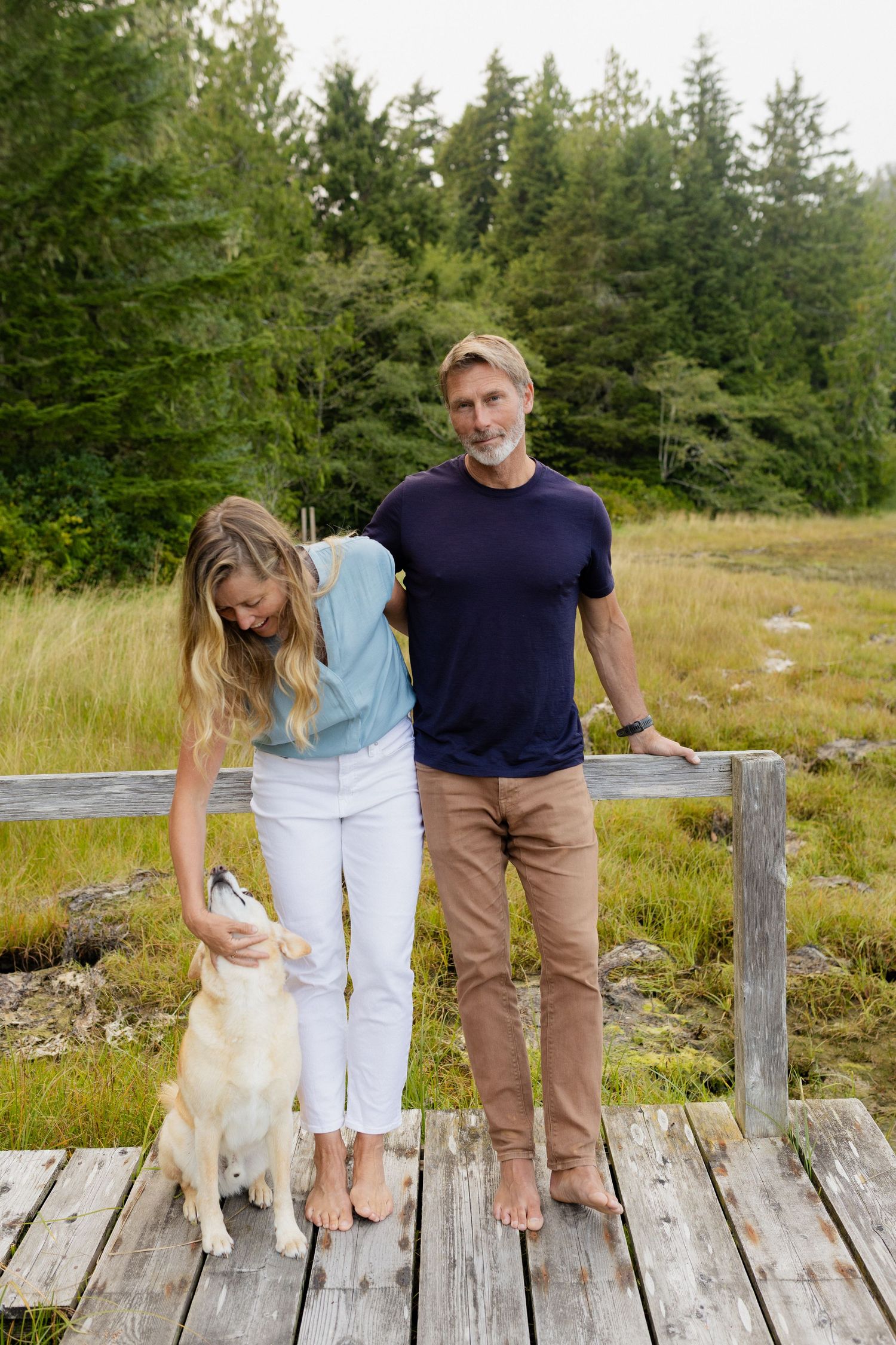 Couple with golden retriever dog on rustic wooden deck surrounded by lush green meadow and forest.