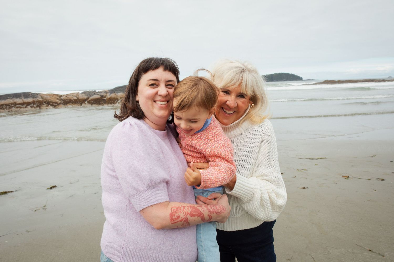 A mother and grandmother smile warmly while holding a young toddler on a sandy beach.