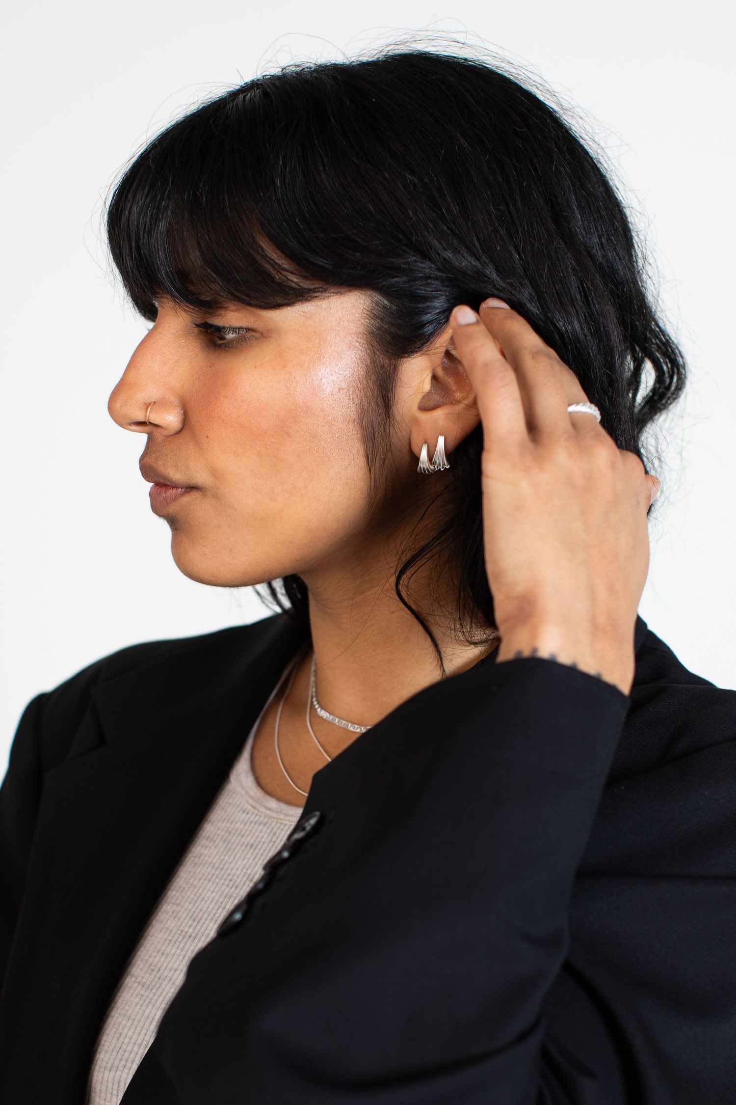 Woman with black hair touching silver hoop earring while wearing black blazer on white background.
