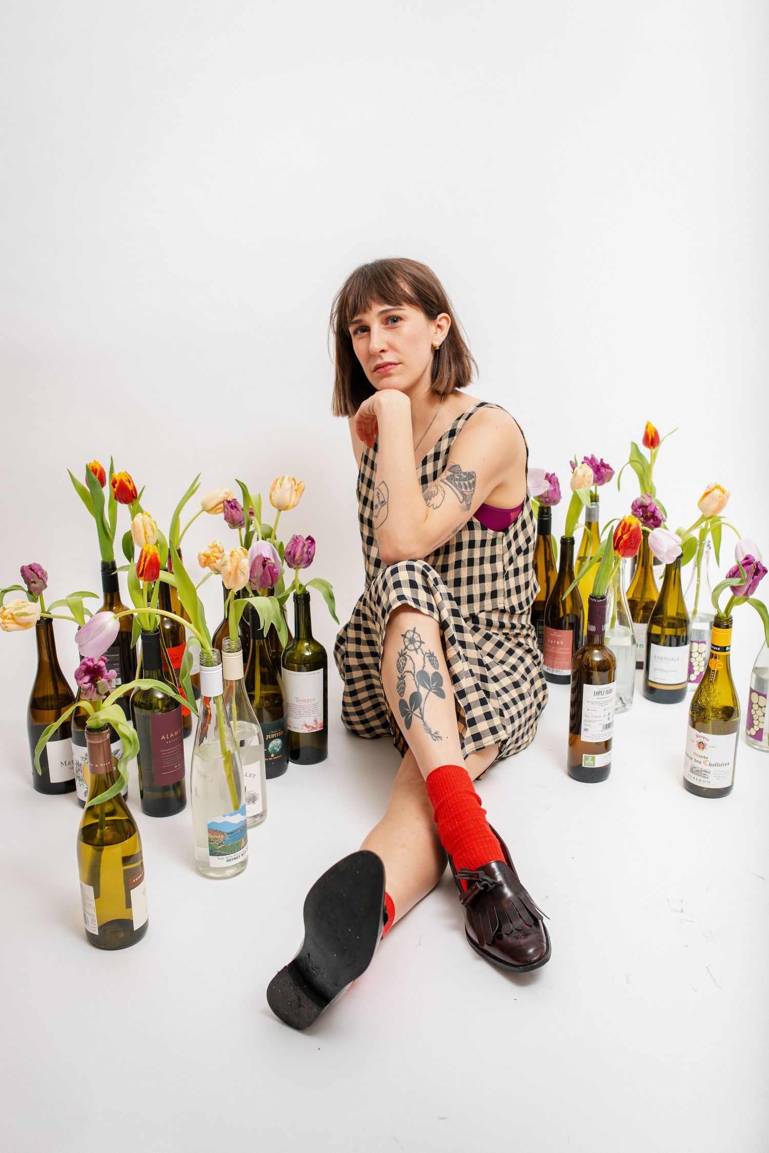 Woman in gingham dress with tattoos sits among wine bottles and colorful tulips on white background.