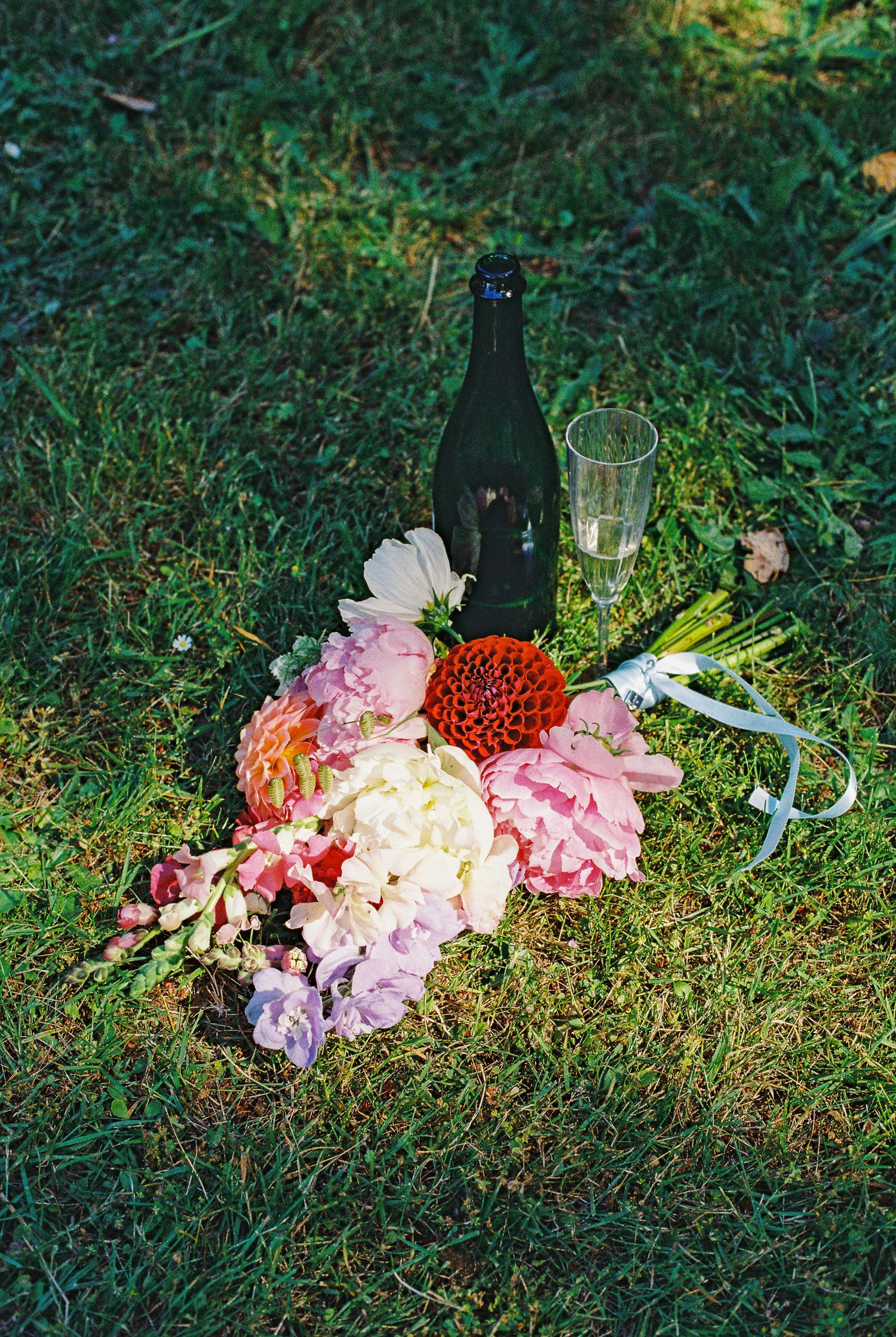 Wedding bouquet of mixed colorful flowers on grass next to a dark green champagne bottle and glass.