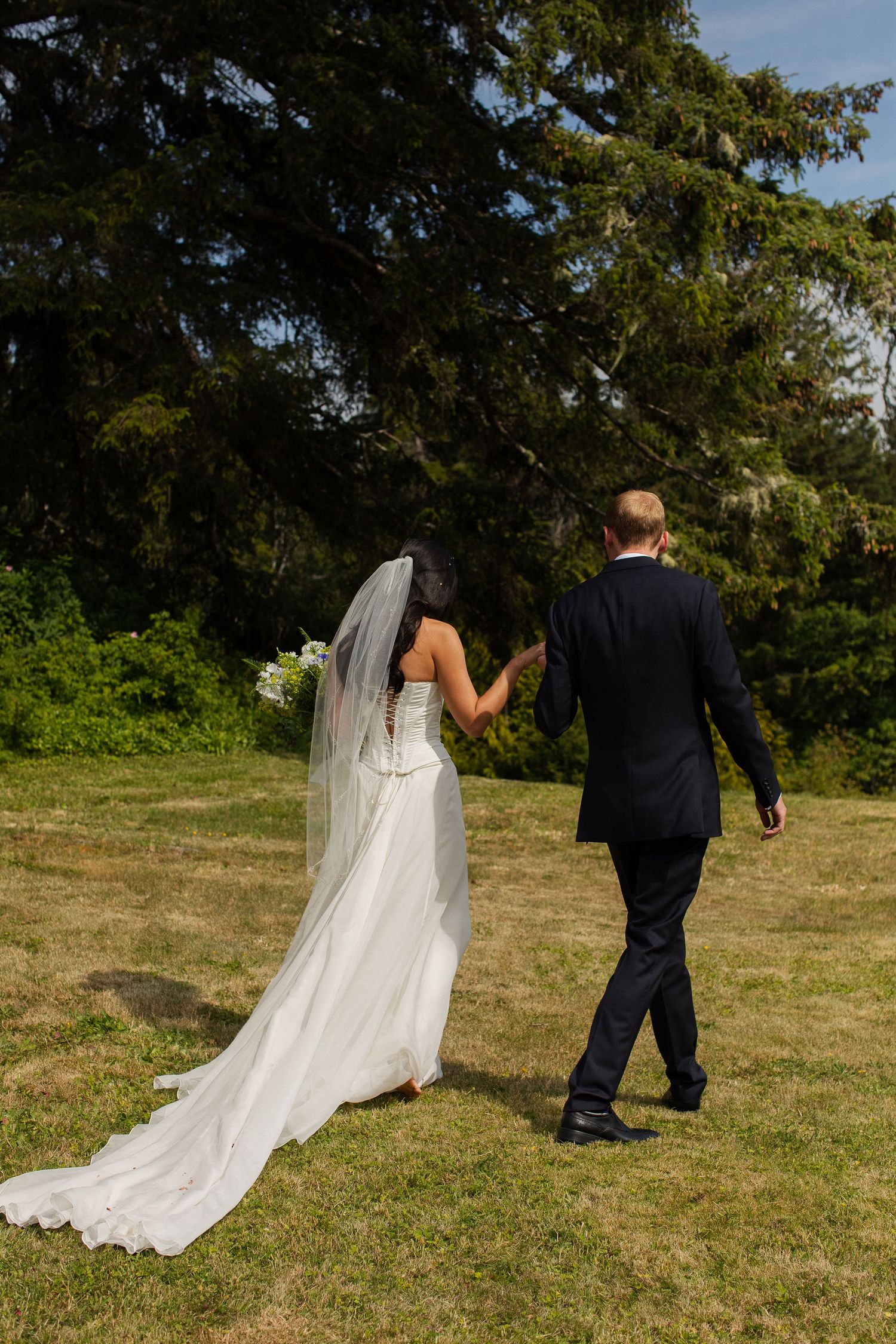 Bride in white gown with long veil and groom in dark suit walking hand in hand across sunny lawn.