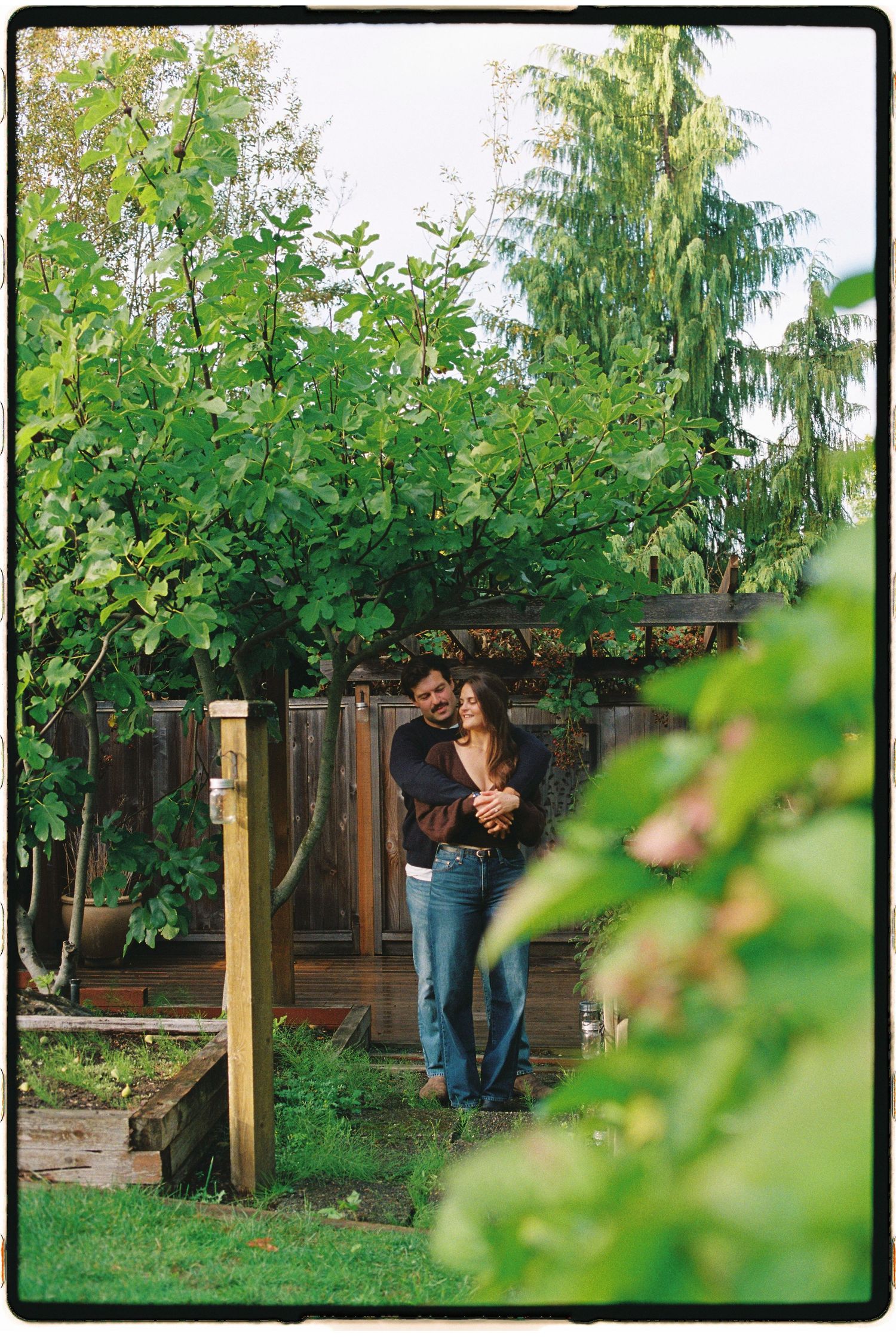 Couple sharing a tender embrace in a lush green garden surrounded by trees and plants.