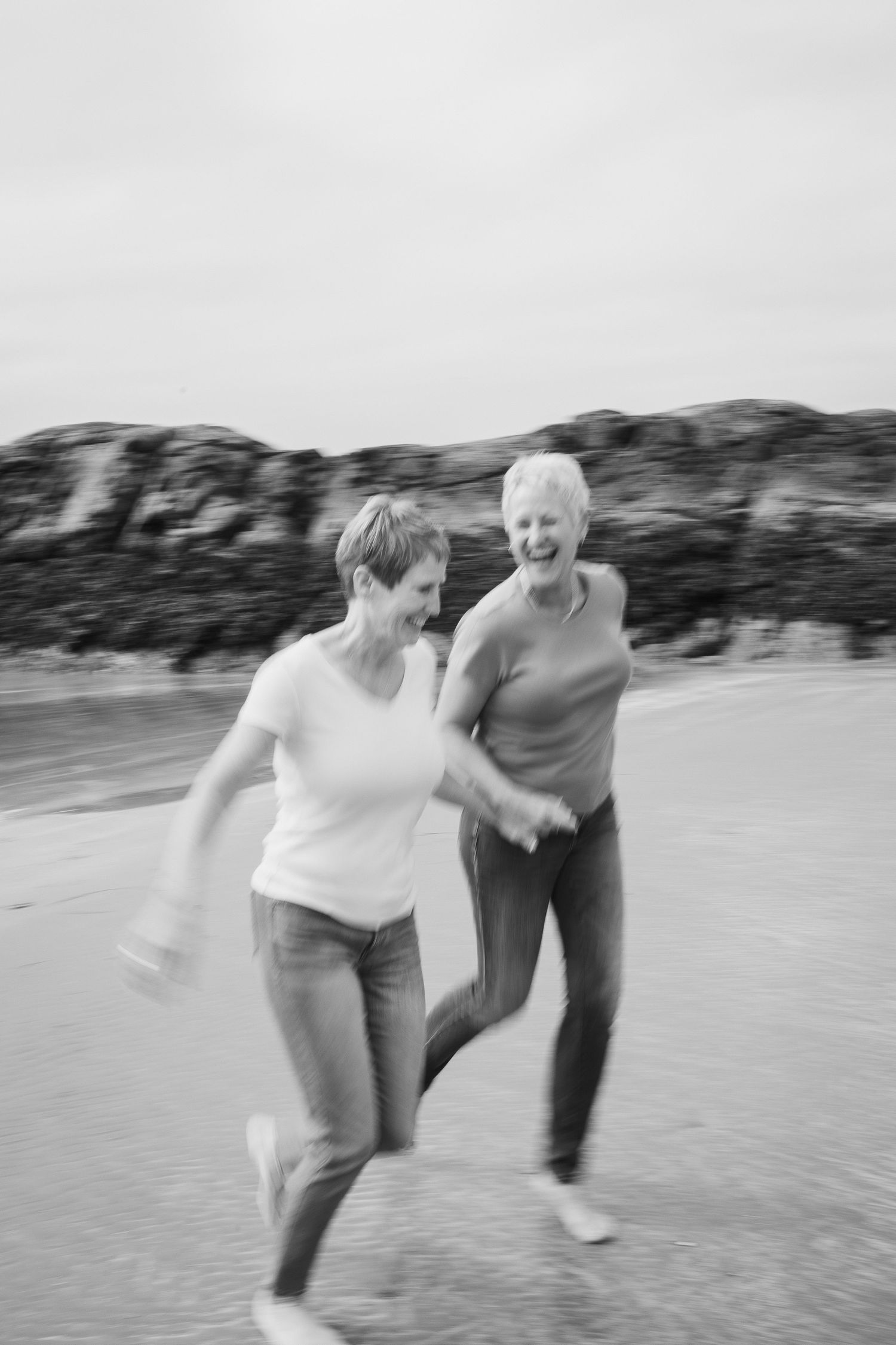 Black and white photo of two women laughing and running playfully along a sandy beach shoreline.