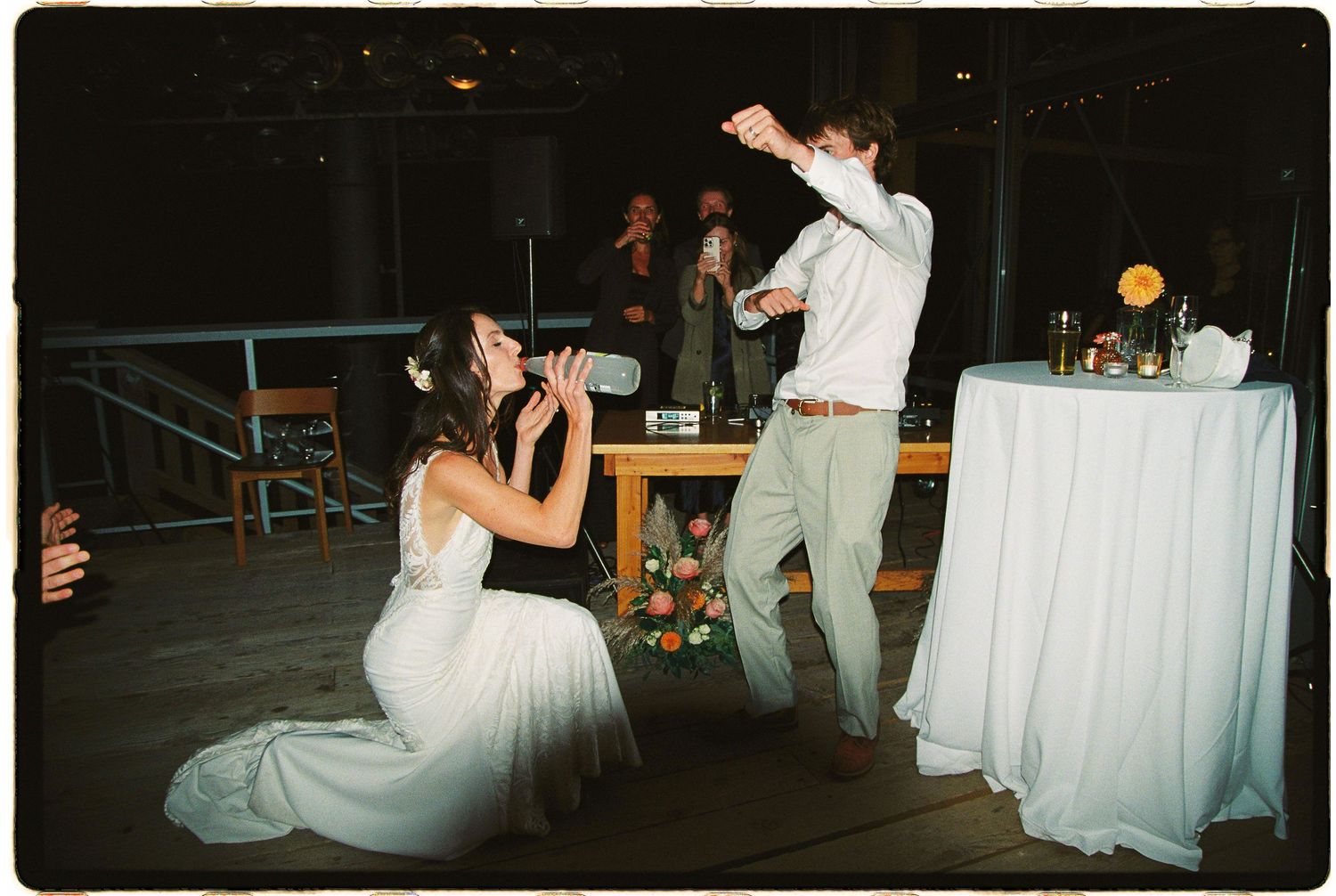 Bride kneeling on dance floor as groom dances at lively indoor wedding reception.