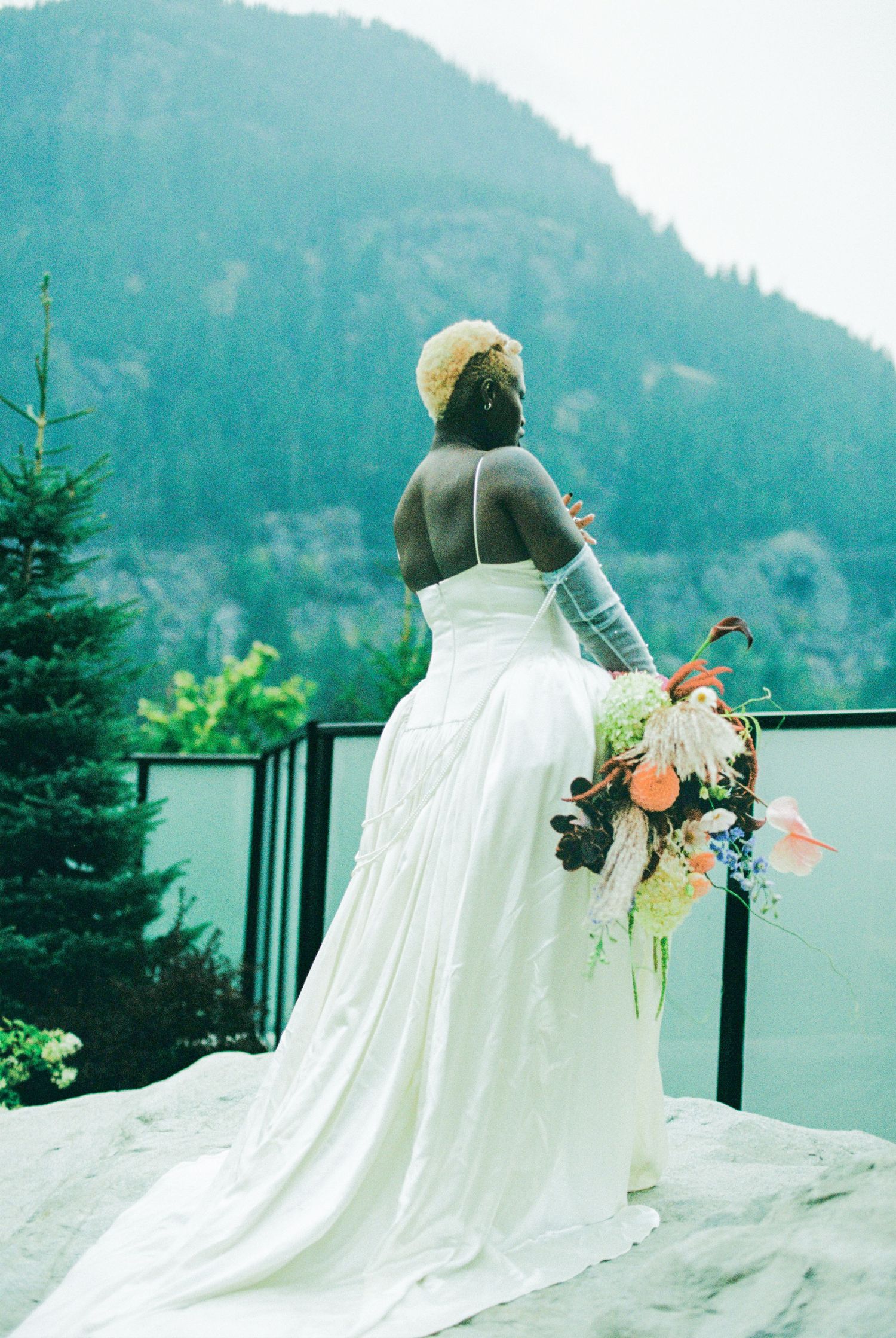 Bride in backless white wedding gown with colorful bouquet gazing at mountain and forest scenery.