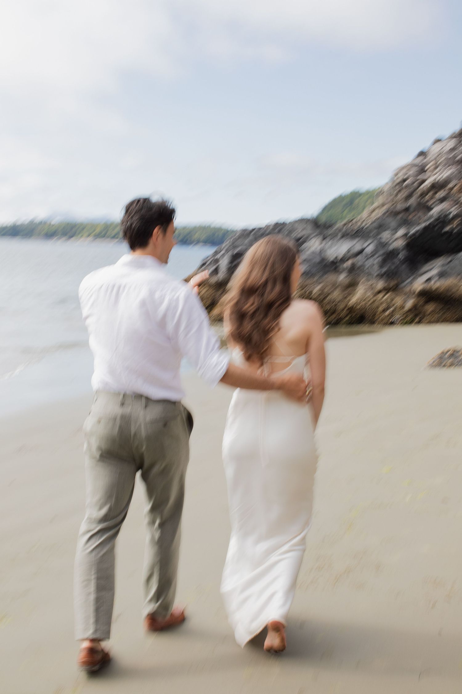 Newlyweds walk barefoot along sandy beach with rocky coastline in a dreamy motion-blur photo.