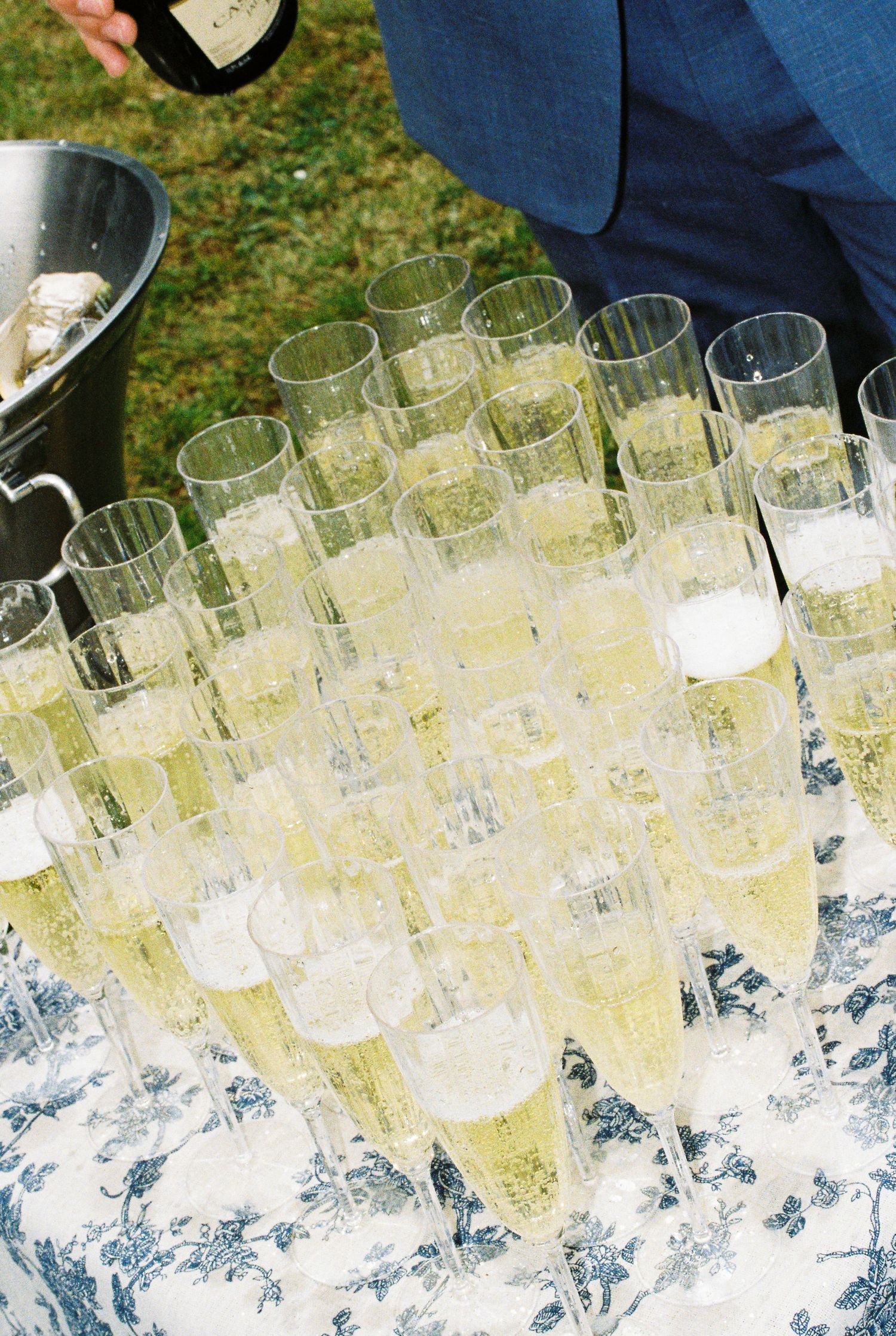 Rows of champagne flutes filled with sparkling wine being poured at an outdoor wedding reception.
