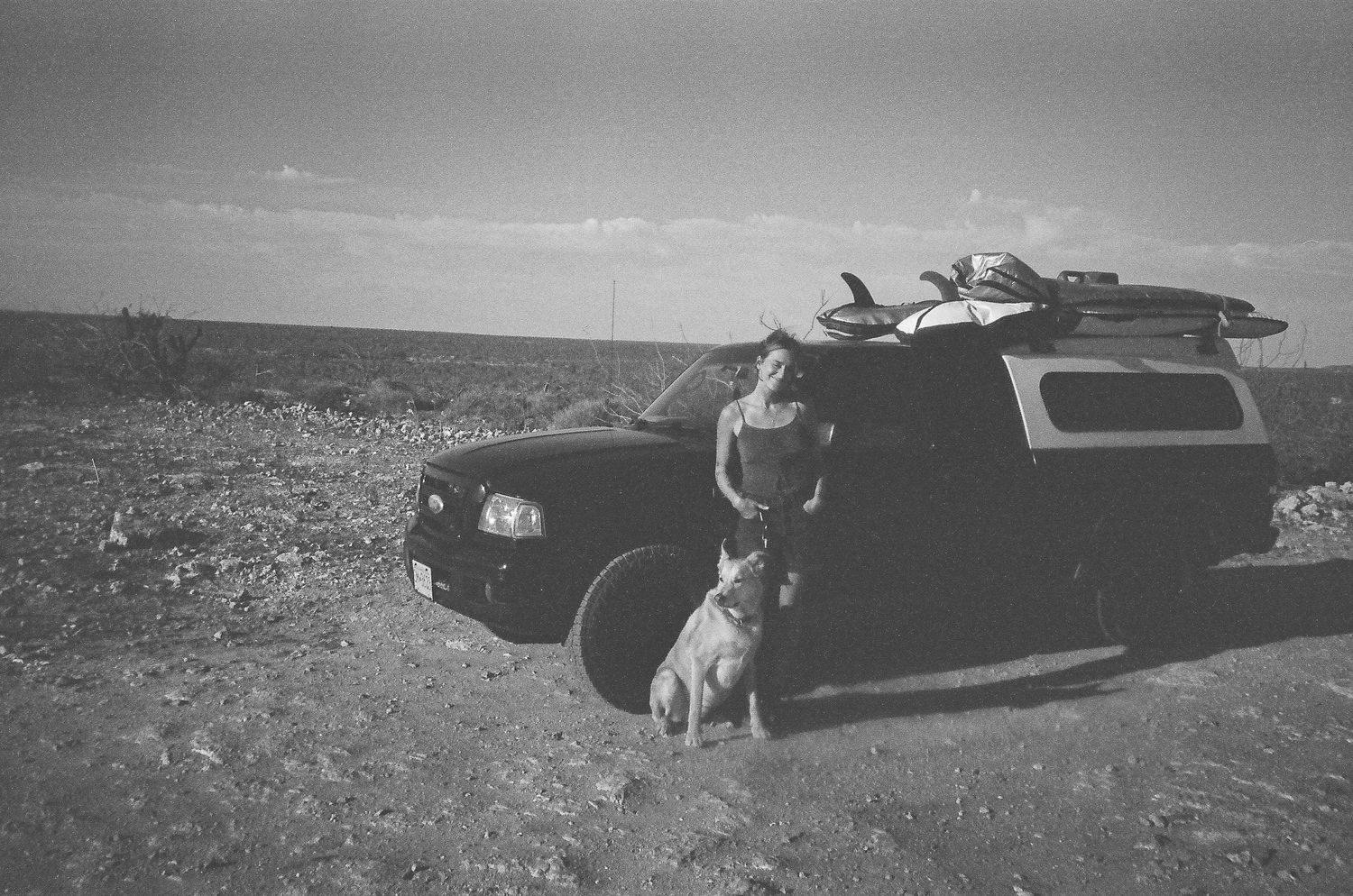 Black and white photo of woman standing beside truck with surfboards and dog in desert landscape.