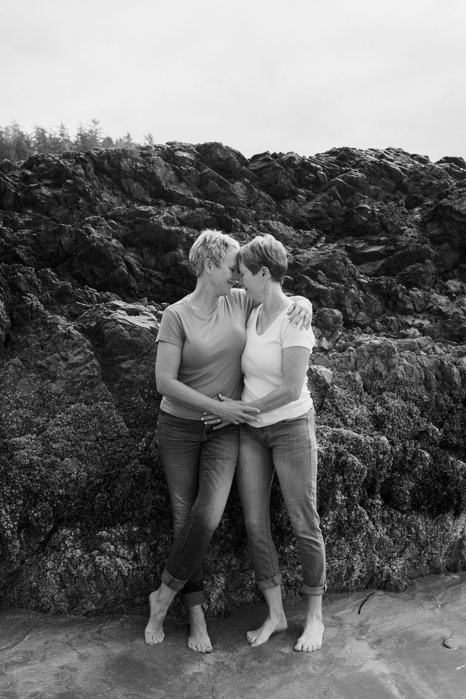 Black and white photo of two women with short hair embracing tenderly against rocky coastal cliffs barefoot.