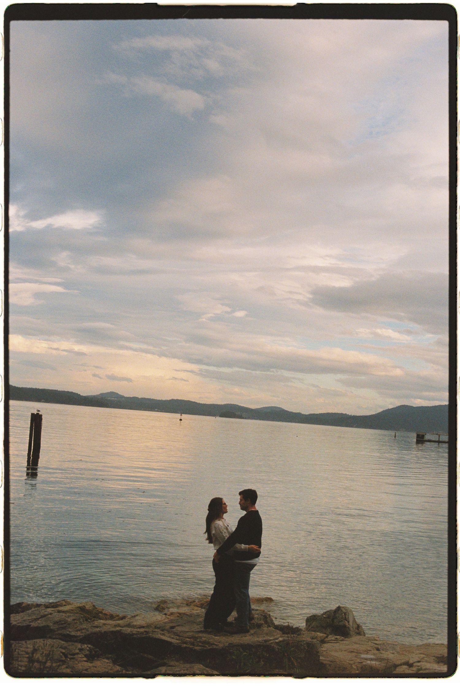 Couple standing close together on rocky shore at sunset with calm lake and cloudy sky.