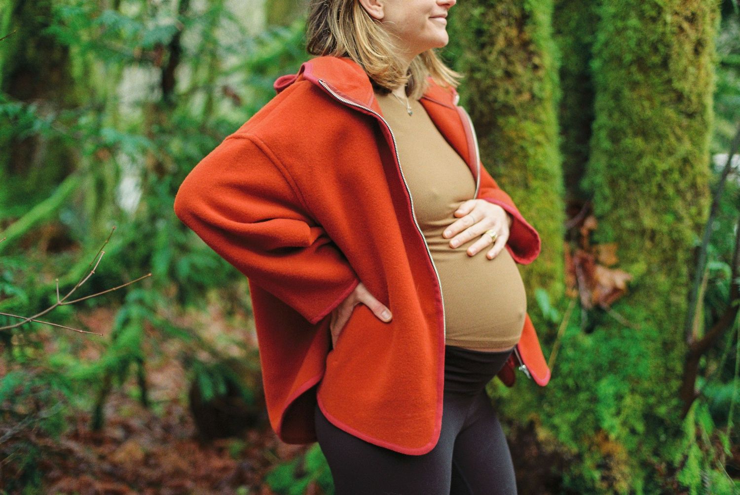 Smiling pregnant woman in red jacket holding baby bump outdoors in lush forest.