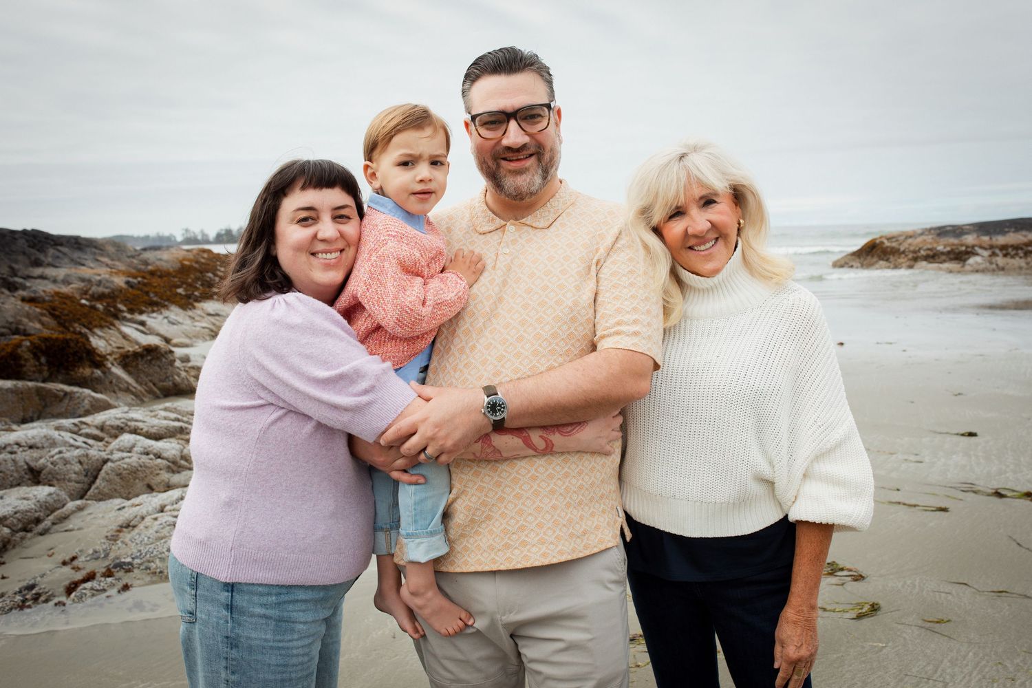 Family of four posing on beach, two adults holding toddler with rocky shoreline behind them.