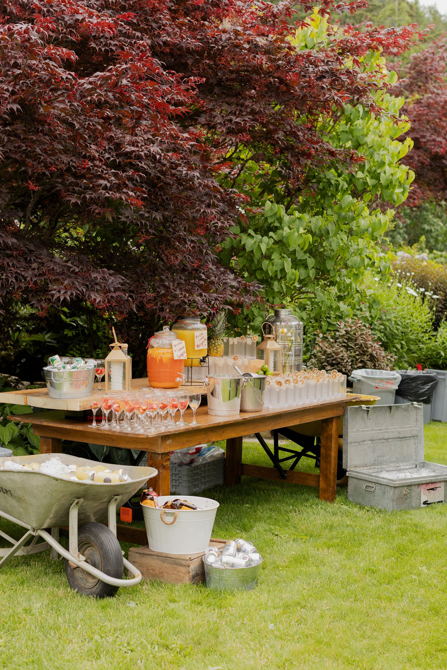 Outdoor wedding beverage station on wooden table with drinks, glassware, and wheelbarrow on lush green lawn.