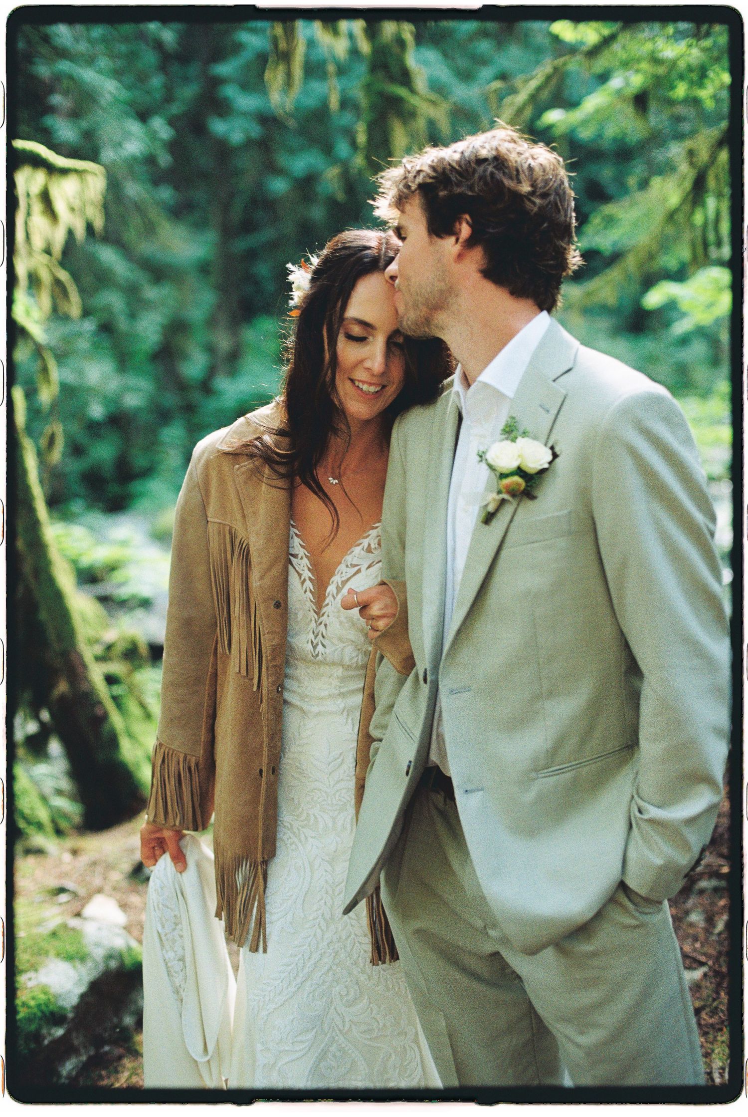 Groom kissing bride on cheek as she smiles wearing fringe jacket in lush green forest.