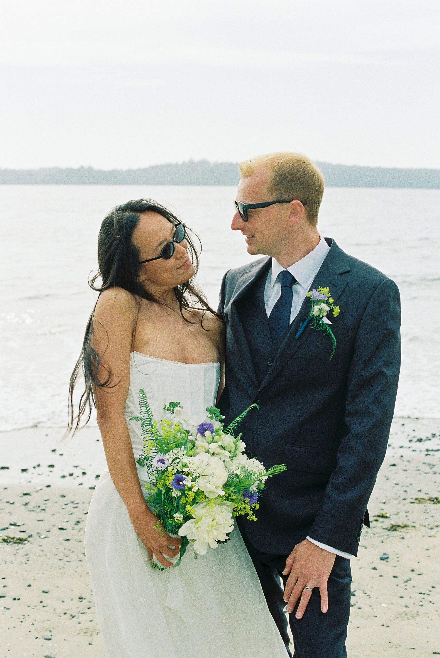 Bride and groom in sunglasses share a tender glance on the beach, groom holding colorful wildflower bouquet.