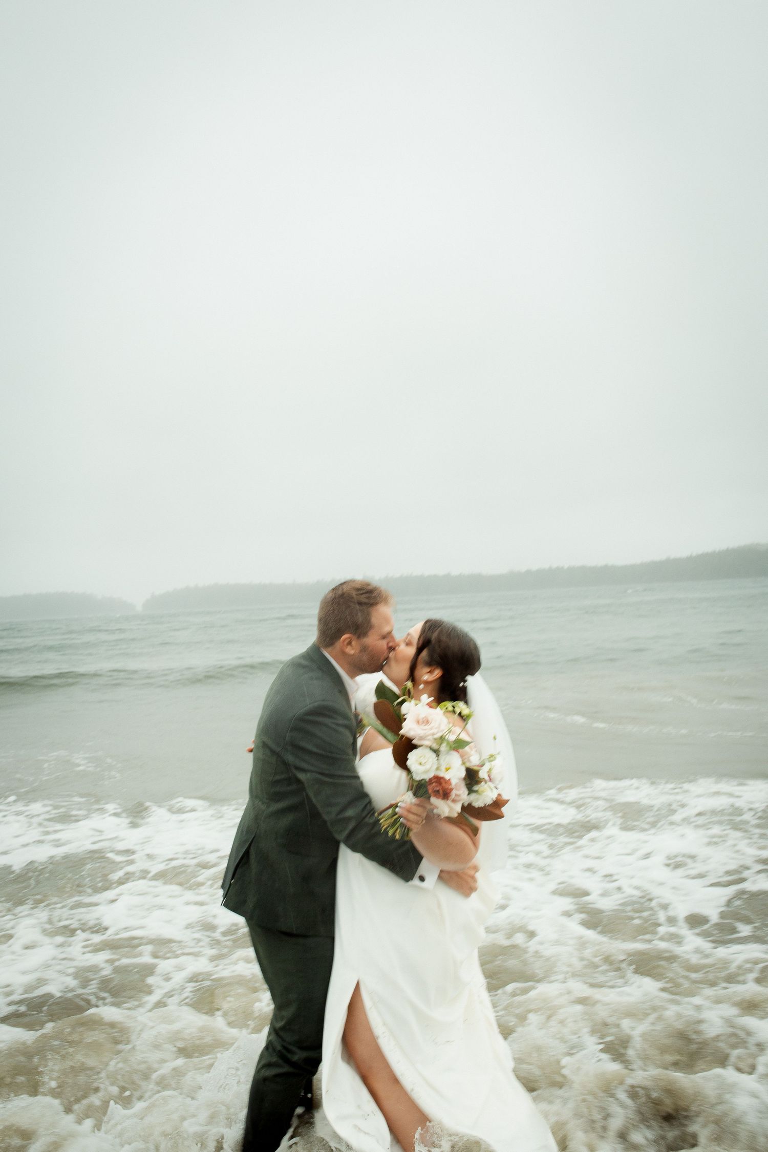 Newlywed couple shares romantic kiss in ocean waves on a misty beach after wedding ceremony.