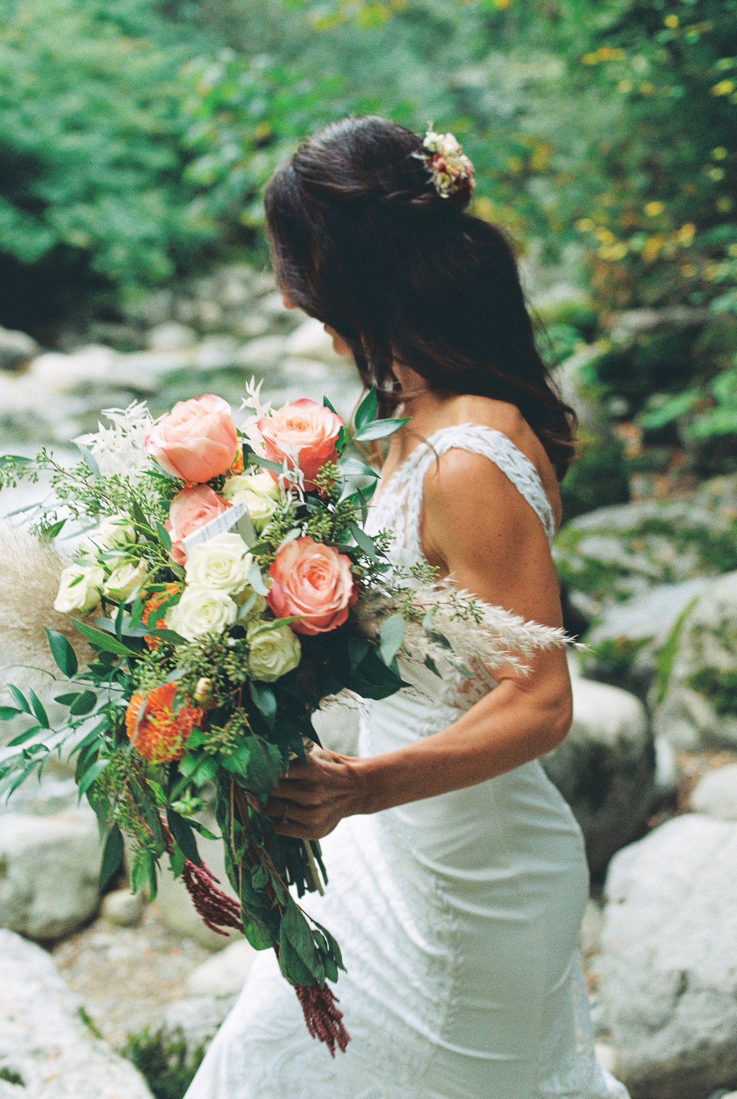 Bride holds lush bouquet of peach roses and greenery in rocky outdoor setting.