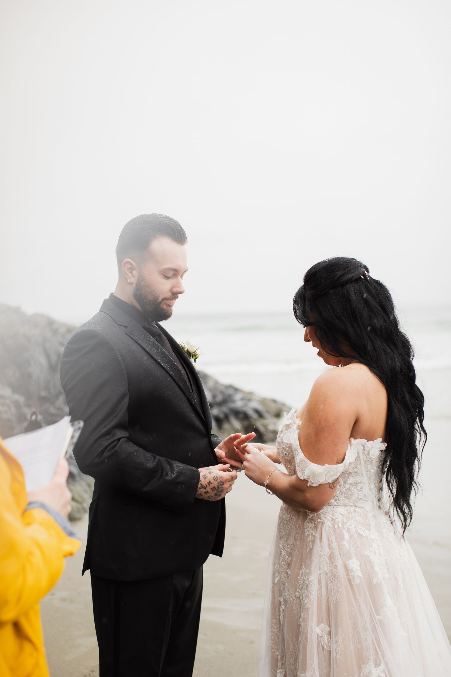 Groom places wedding ring on bride's finger during romantic beach ceremony on overcast day.