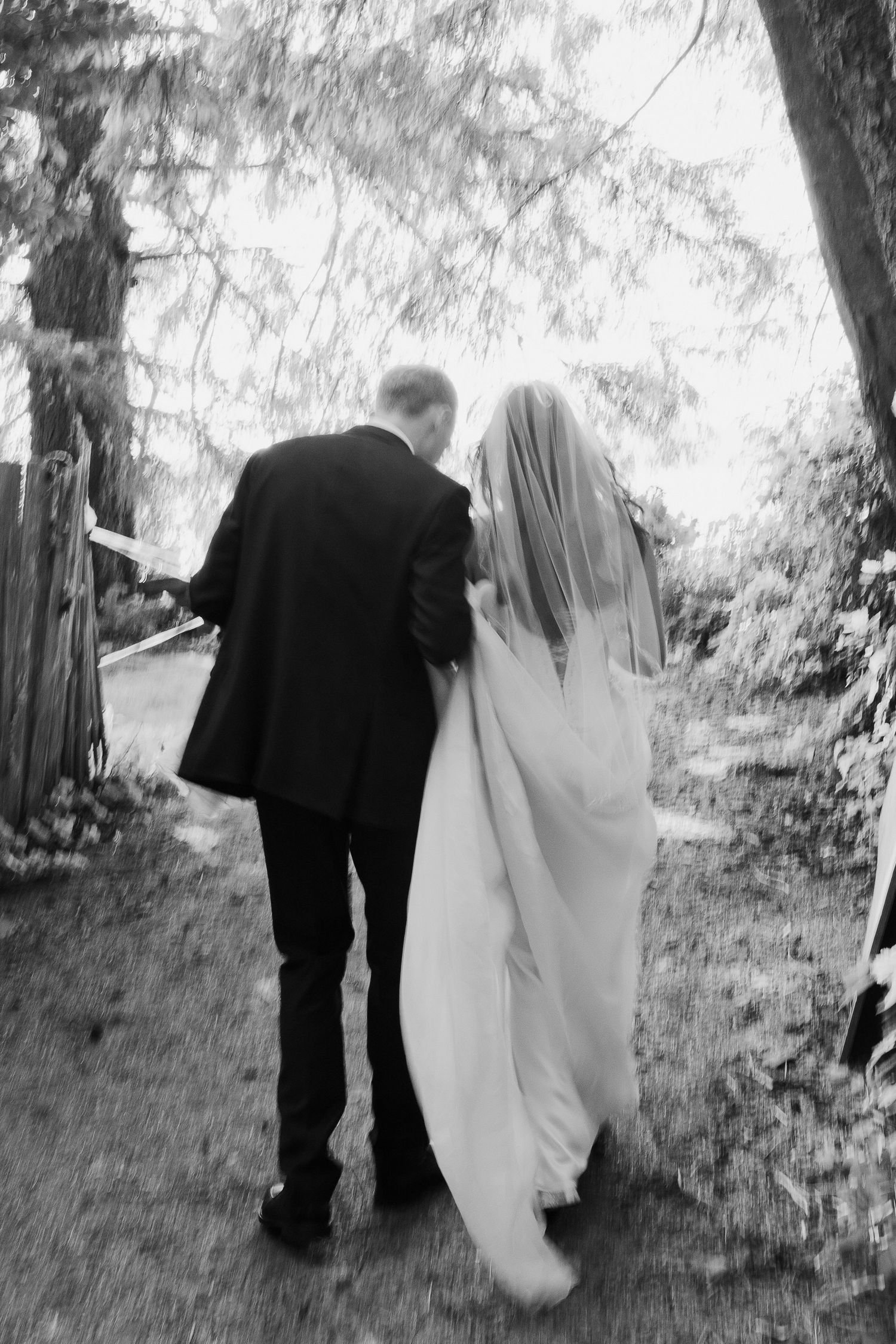 Black and white photo of bride and groom walking hand in hand through a tree-lined garden path.