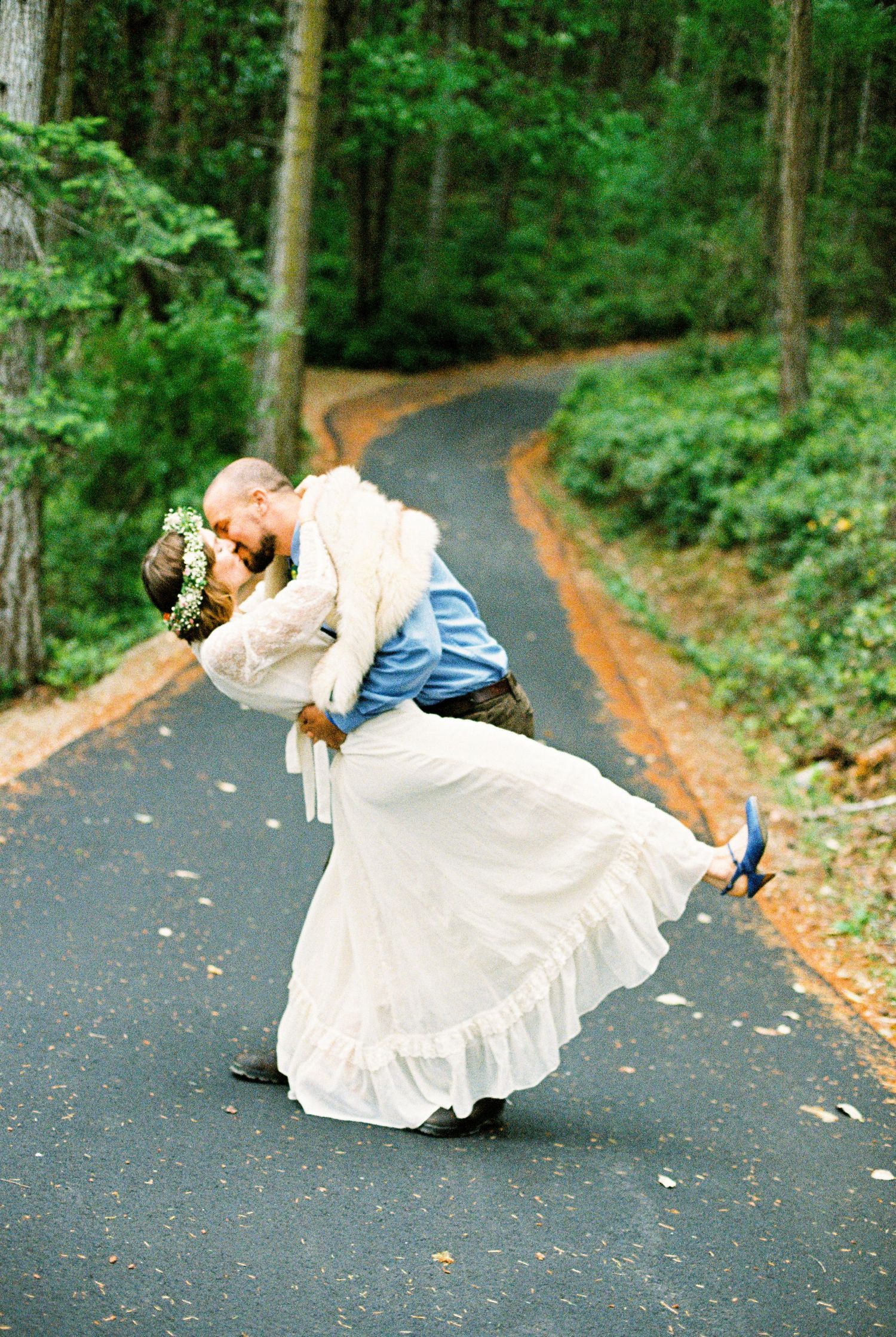 Groom dipping bride in white gown for a romantic kiss on a forest road.