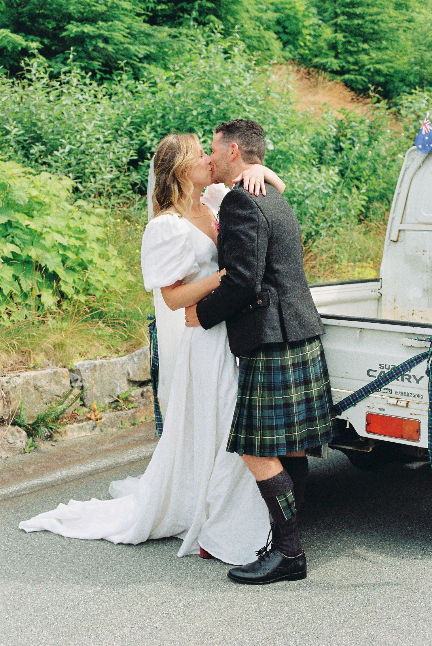 Bride and groom kissing beside vintage white truck on a lush green country road.