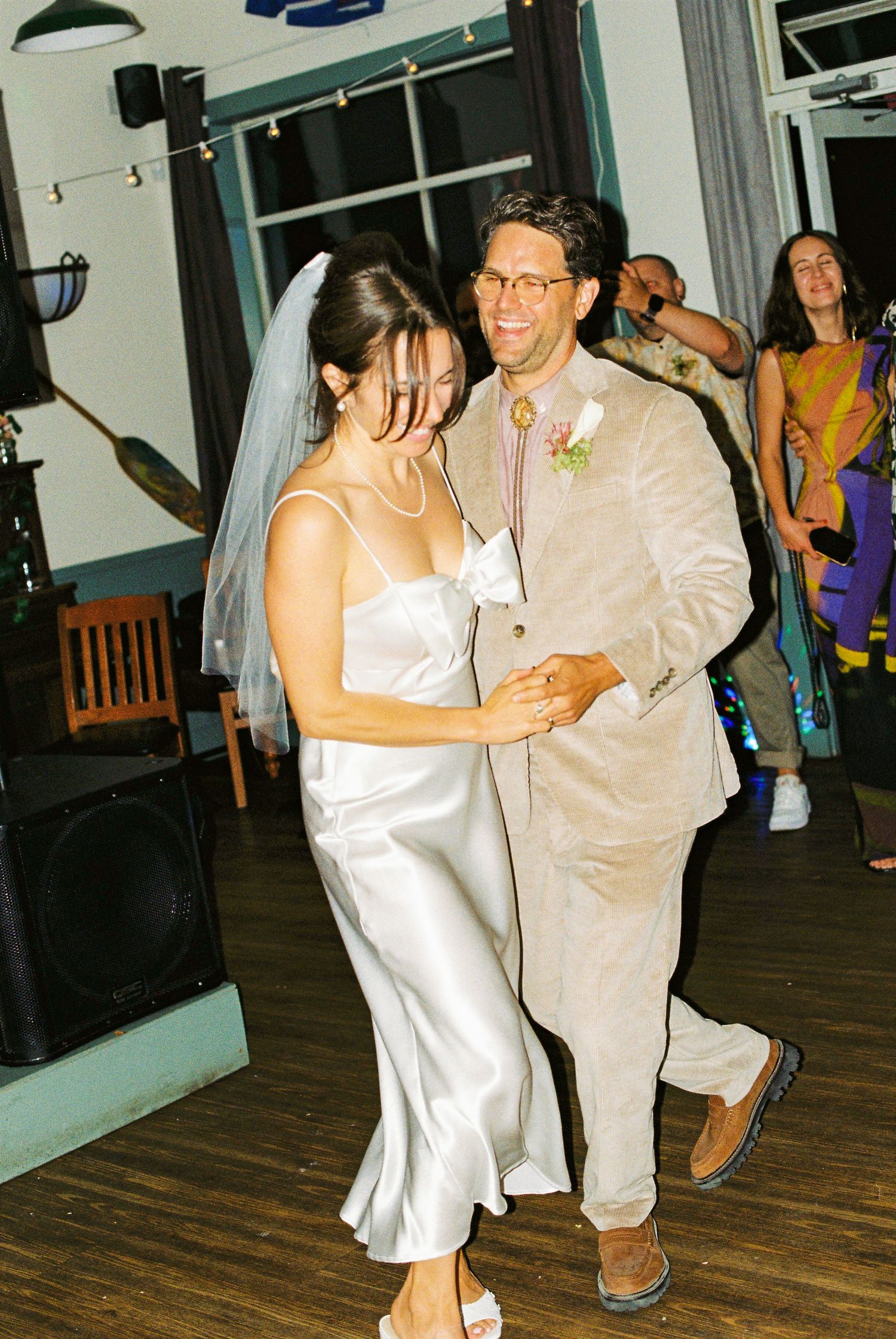 Bride in white satin slip dress and veil dances with groom in linen suit at indoor wedding reception.