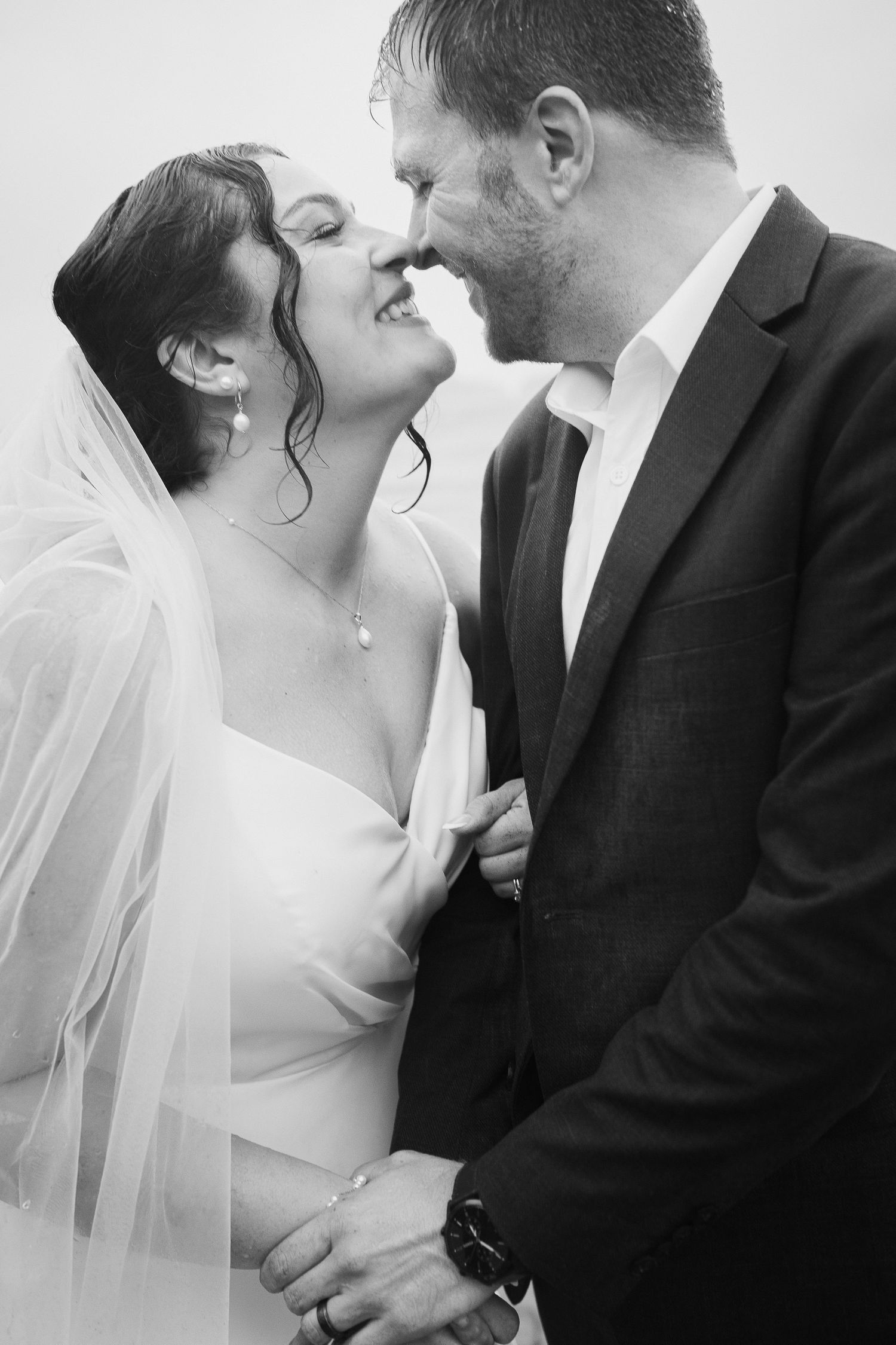 Black and white photo of bride and groom smiling and almost kissing, bride wearing veil and elegant gown.