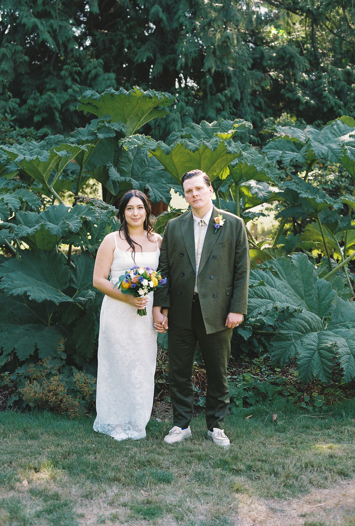 Bride and groom holding hands posing before large tropical plants at outdoor garden wedding.