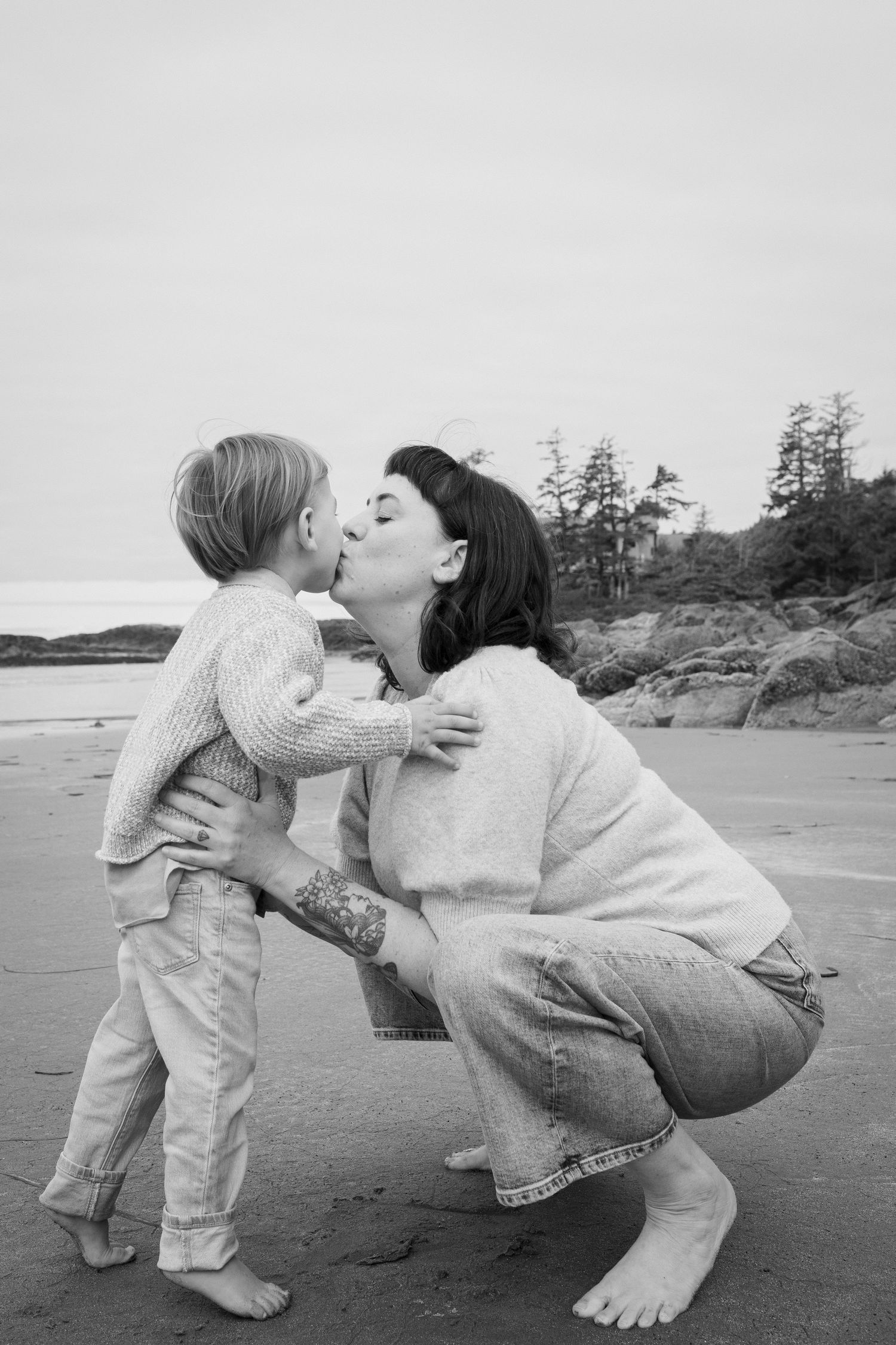 A mother crouches to kiss her young child on a sandy beach in a tender black and white portrait.