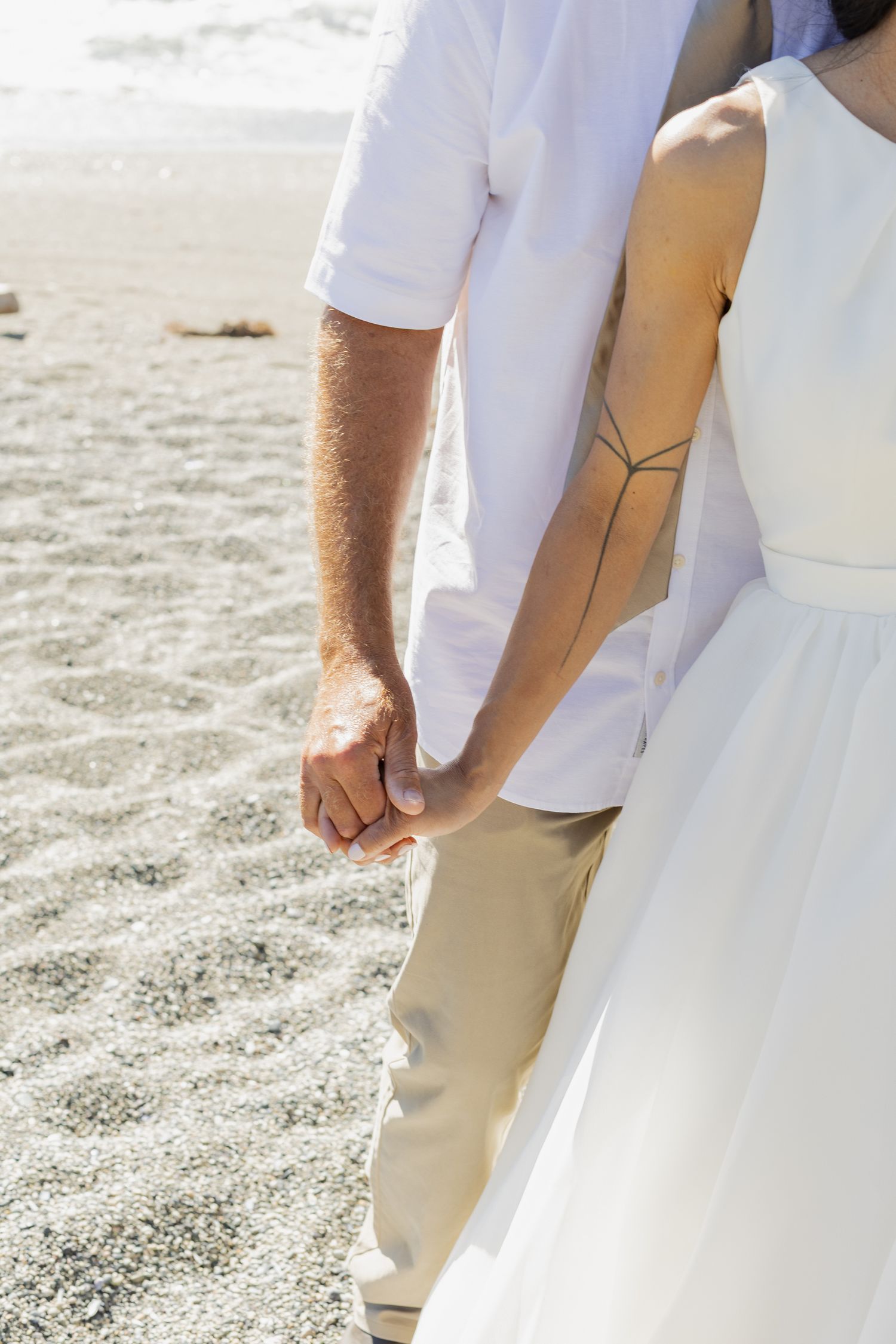 Close-up of bride with back tattoo and groom holding hands on sandy beach wedding day.