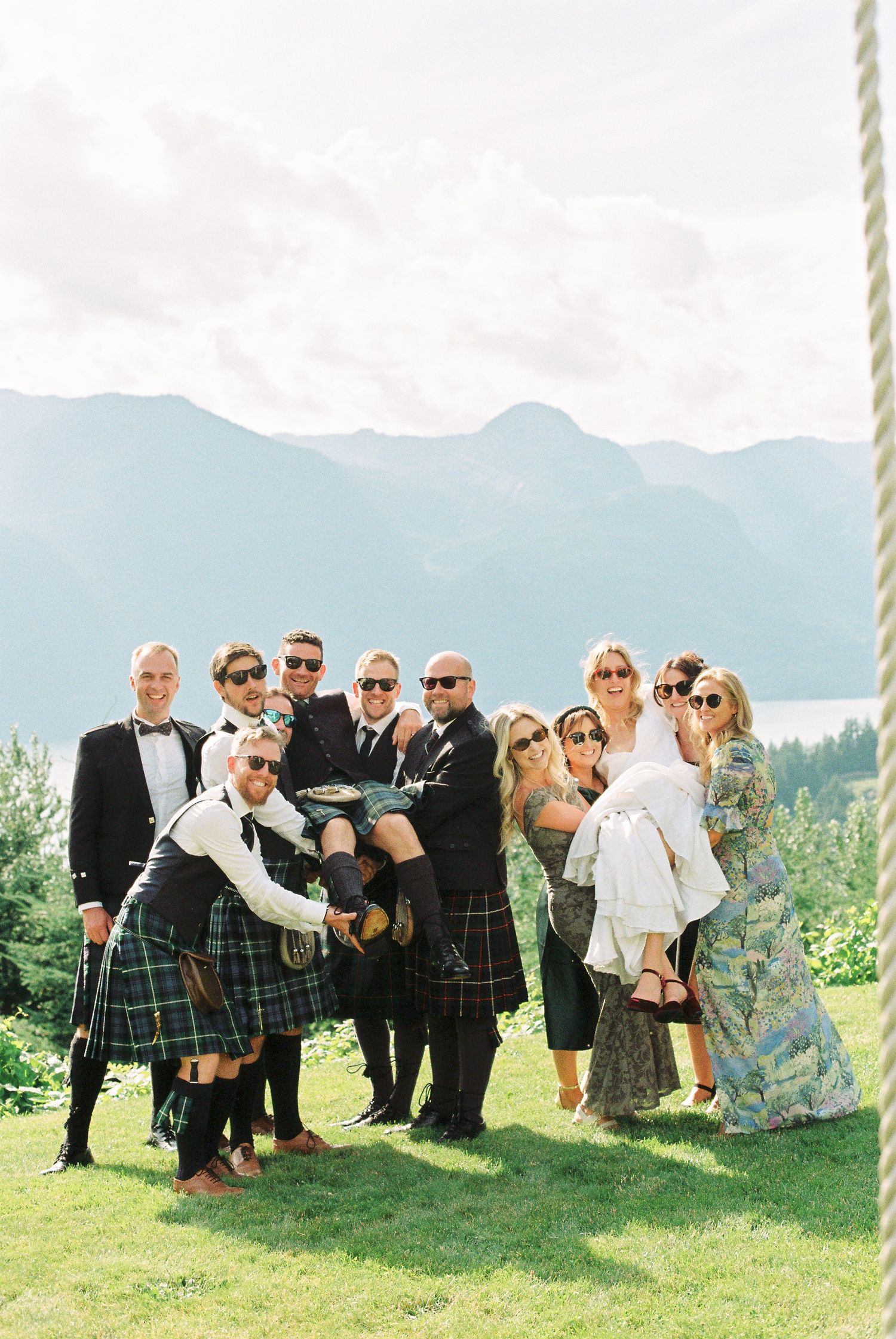 Wedding party in kilts and formal wear posing outdoors with mountain lake backdrop.