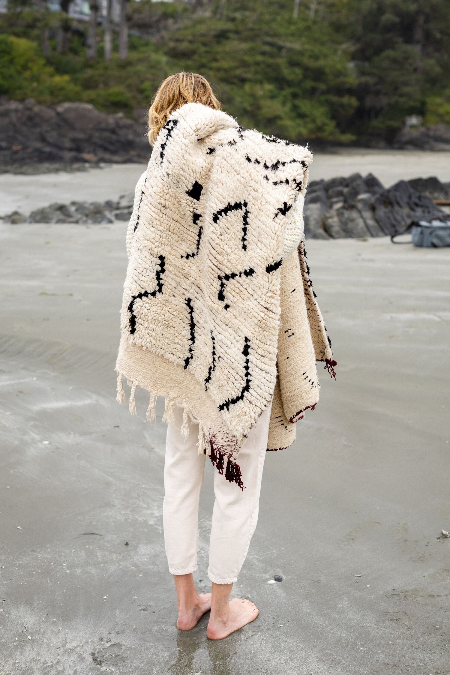 Woman wrapped in cream Berber blanket with black pattern stands barefoot on sandy beach.